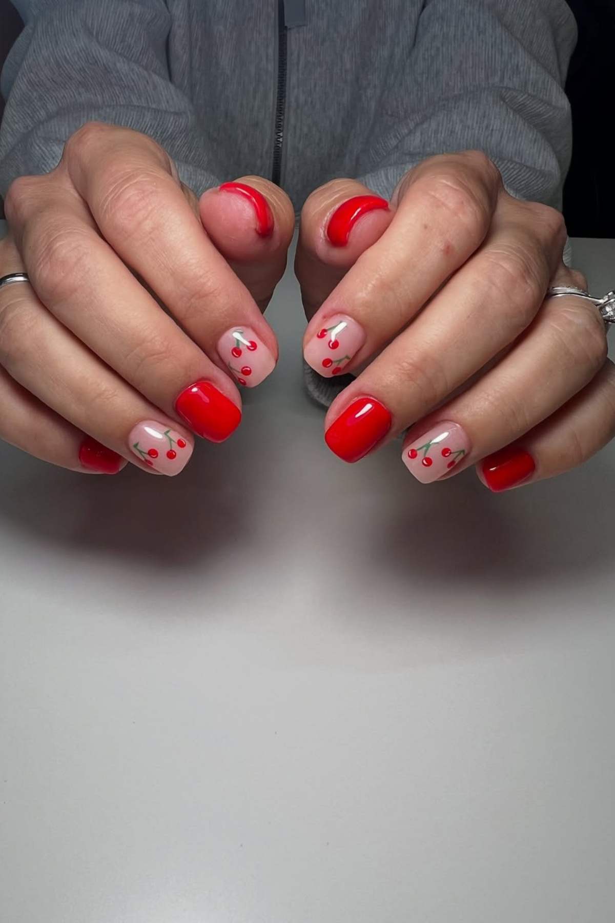 Hands with short, square-shaped nails painted red, with some nails featuring a light base and small red cherry designs. Person wears two rings and a grey zip-up top.