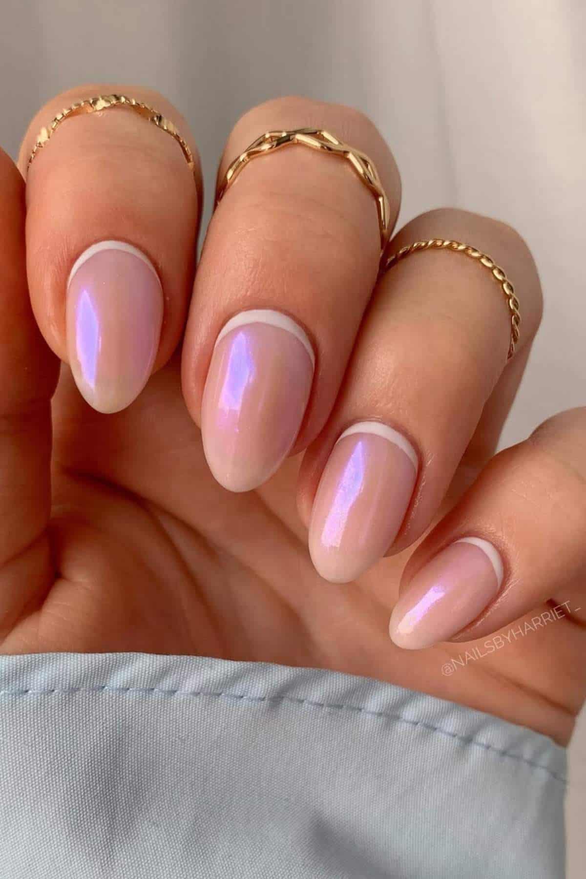 A hand with almond-shaped nails featuring a pink, iridescent manicure and gold rings on each finger.