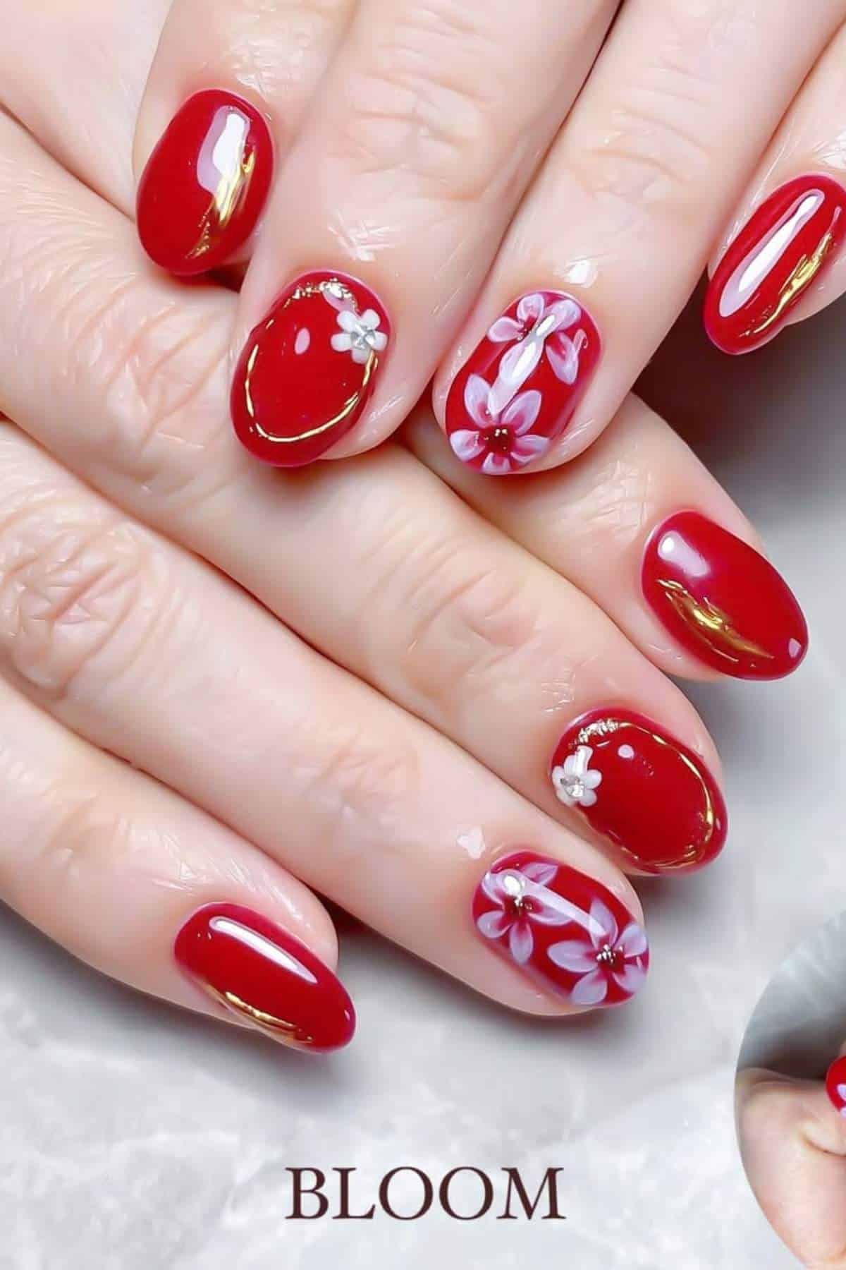 Hands with glossy red nail polish; some nails feature white and pink flower designs and gold accents. The word "BLOOM" appears at the bottom of the image.