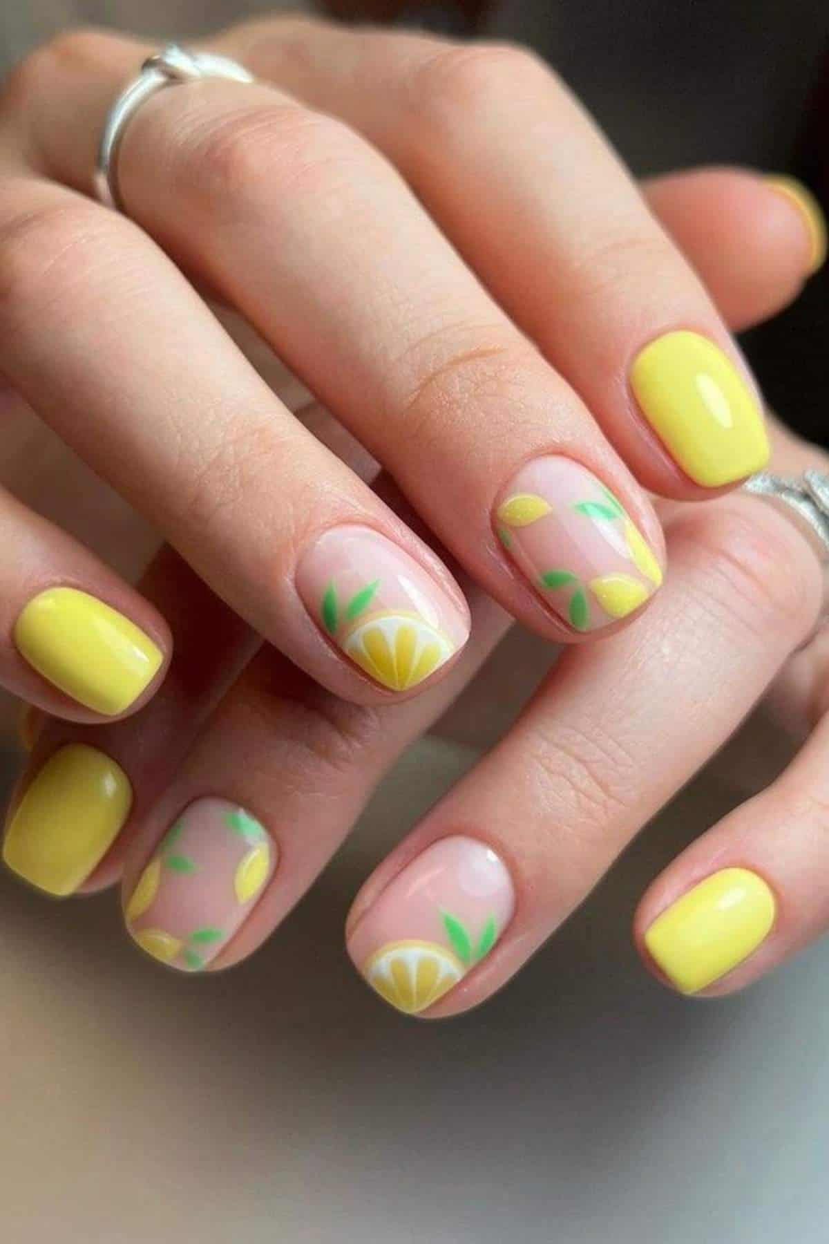 Hands with short, yellow-painted nails; some nails feature lemon and leaf designs on a nude background. The person wears silver rings.