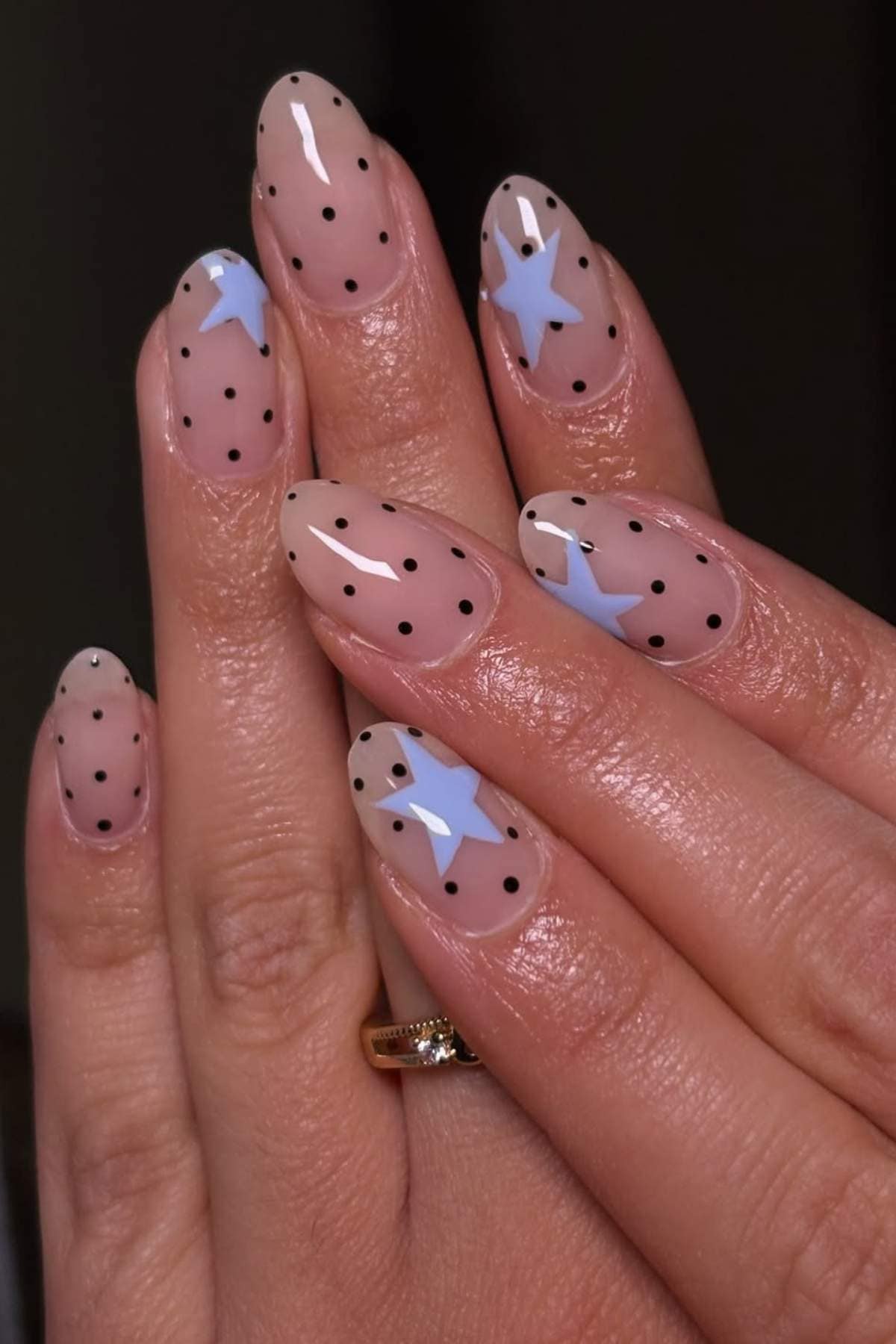 A hand with almond-shaped nails featuring a nude base, black polka dots, and blue star accents on two nails.