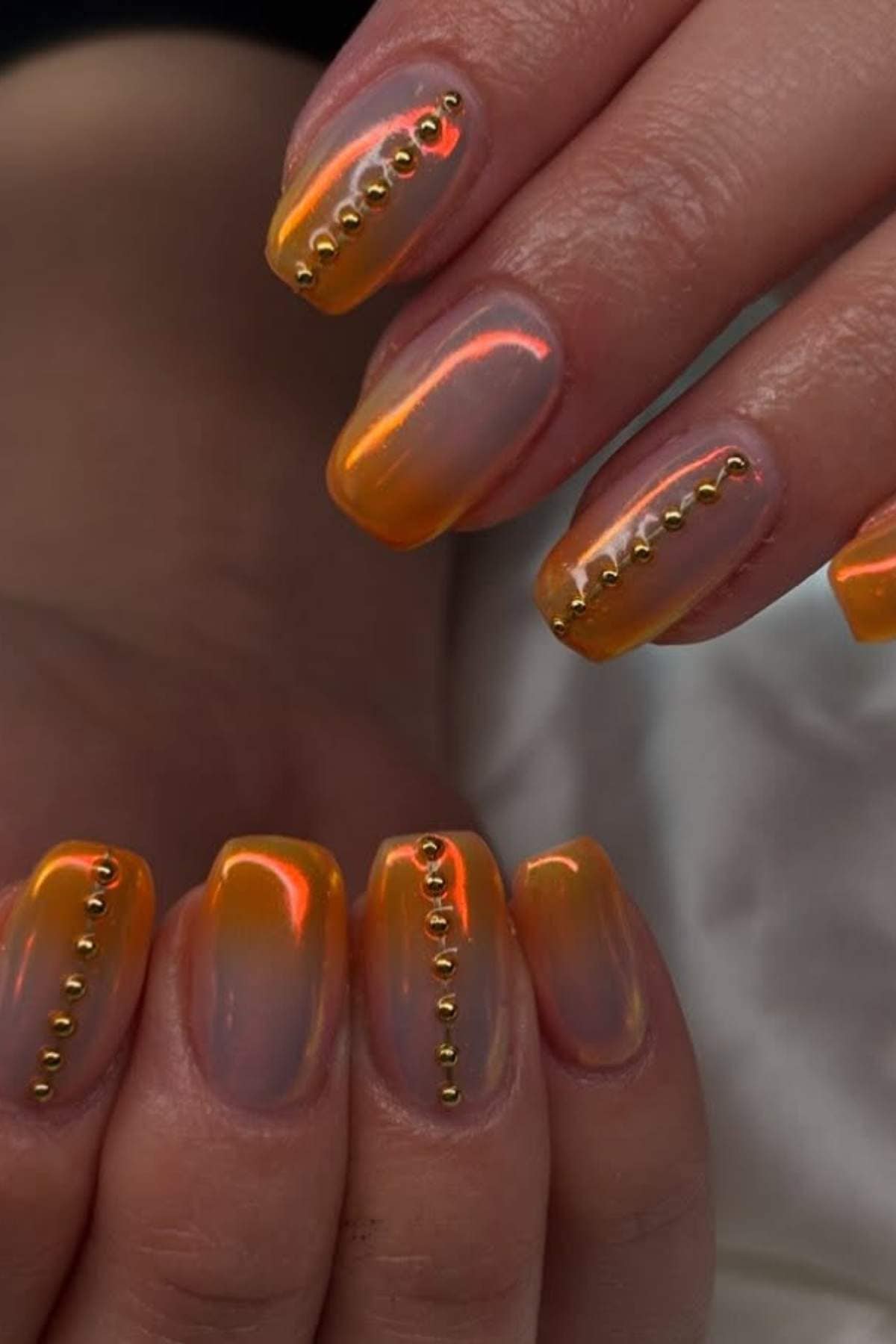 Hands with orange, iridescent nails featuring a vertical line of small gold studs on each nail.