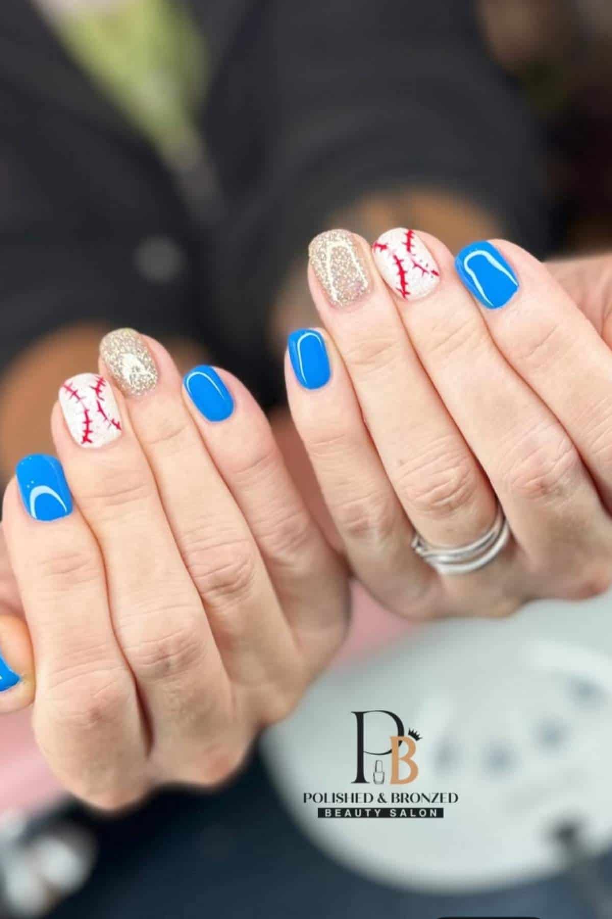 Hands with a manicure featuring blue nails, gold glitter nails, and white nails with red stitching resembling a baseball. The Polished & Bronzed Beauty Salon logo is visible.