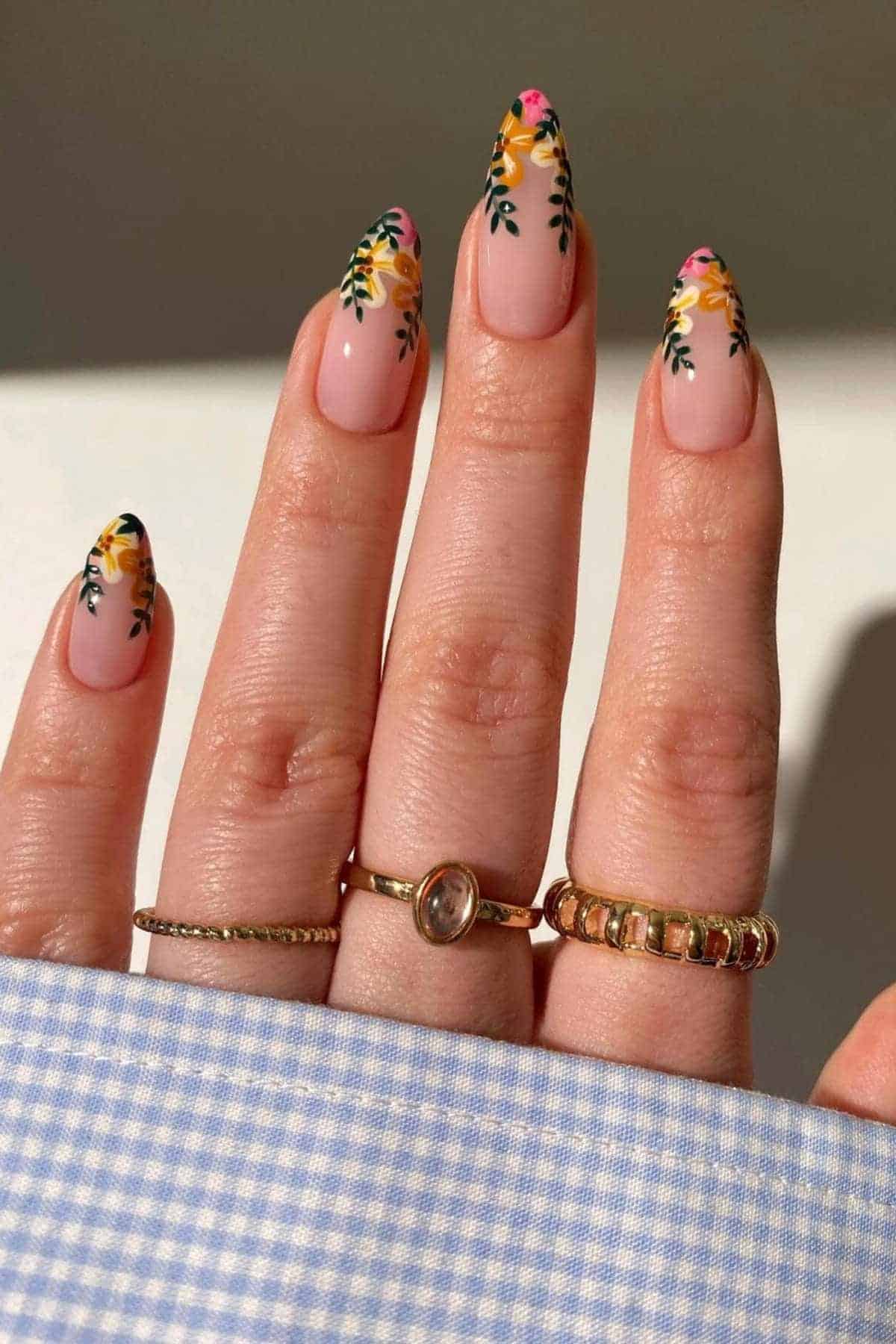A hand with long almond-shaped nails featuring yellow and pink floral nail art, wearing three gold rings, resting on blue gingham fabric.