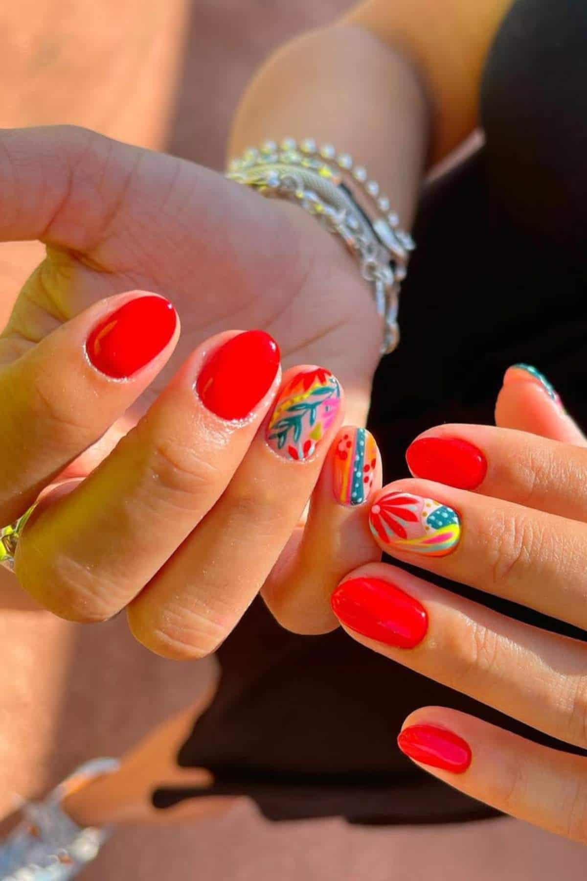 A person displays manicured hands with bright red nail polish and colorful, intricate designs on the ring and pinky fingernails.