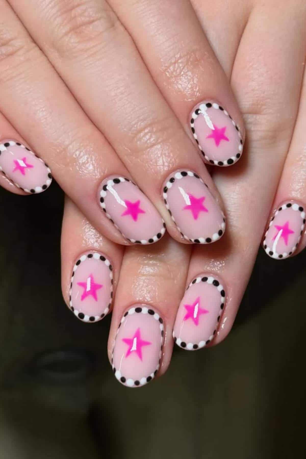 Close-up of hands with nude nail polish, each nail featuring a pink star in the center and black polka dots outlining the nail edges.