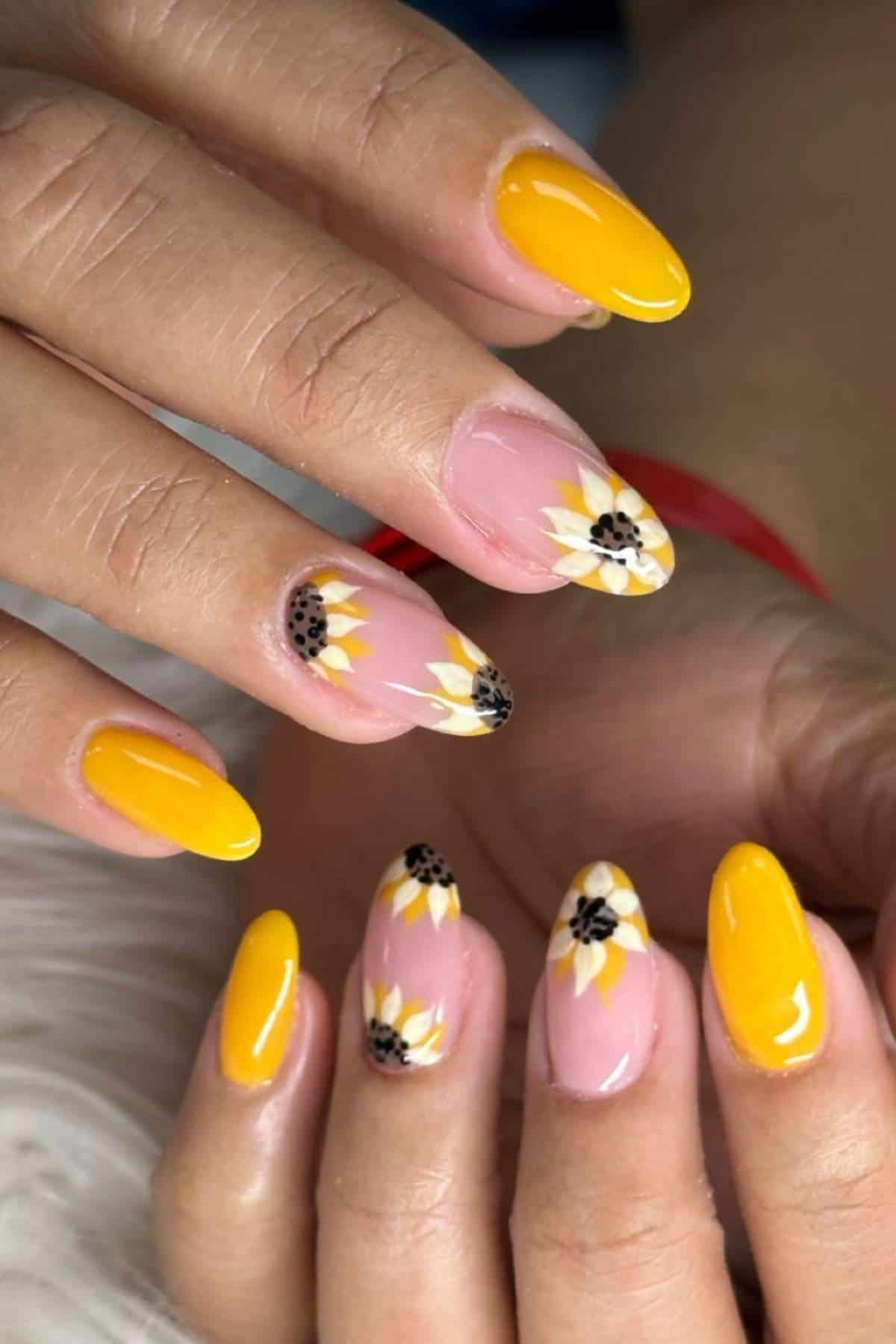 Close-up of hands with almond-shaped nails, some painted yellow and others decorated with sunflower designs on a nude base.
