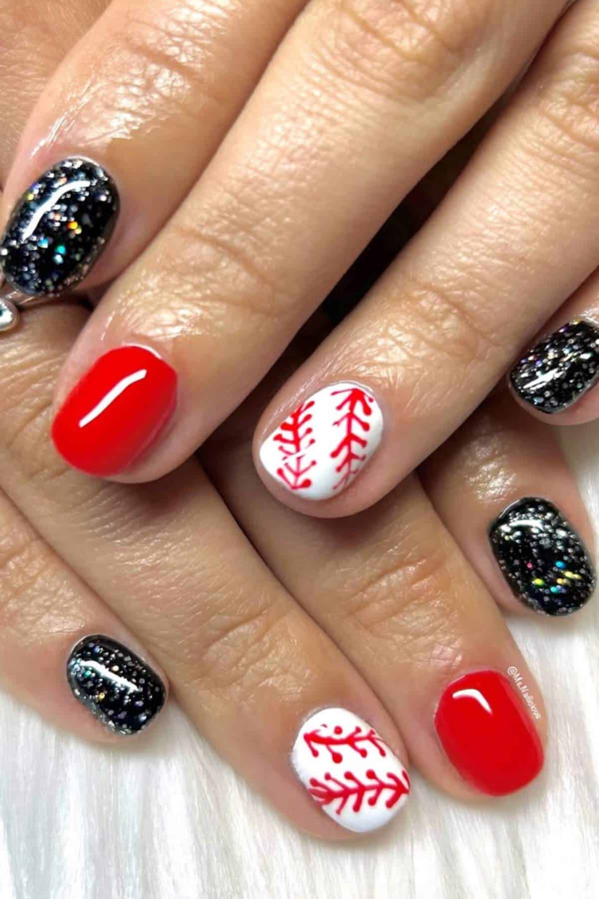 A hand with manicured nails featuring black glitter, solid red polish, and a white nail with red baseball stitch design.