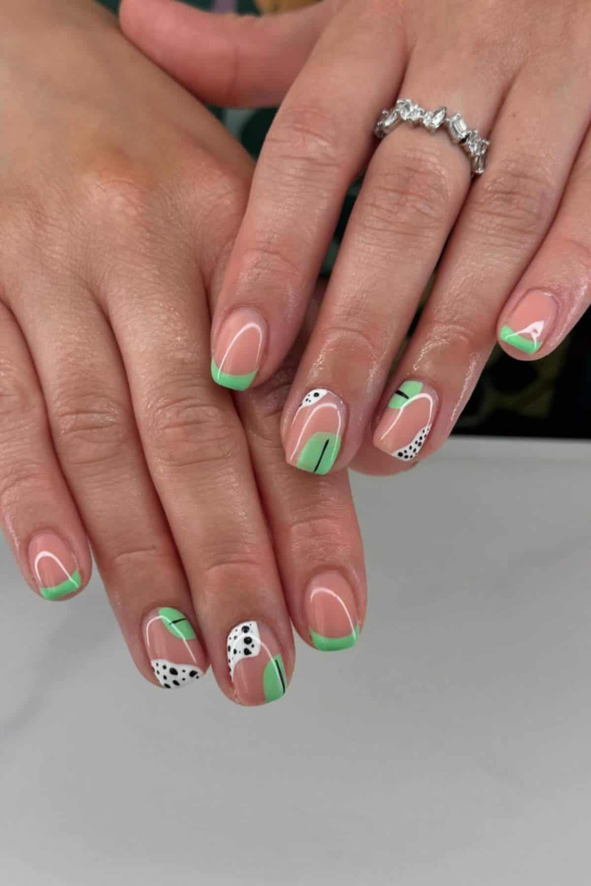 Hands with manicured nails featuring green, white, and black abstract designs and a silver ring on the ring finger.