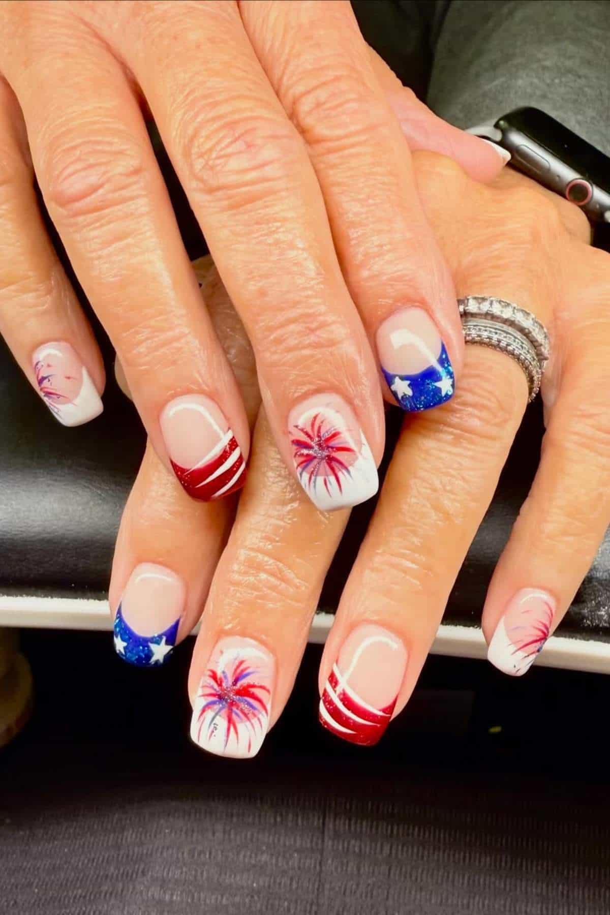 French tip nails with red, white, and blue patriotic designs, including fireworks and stars.