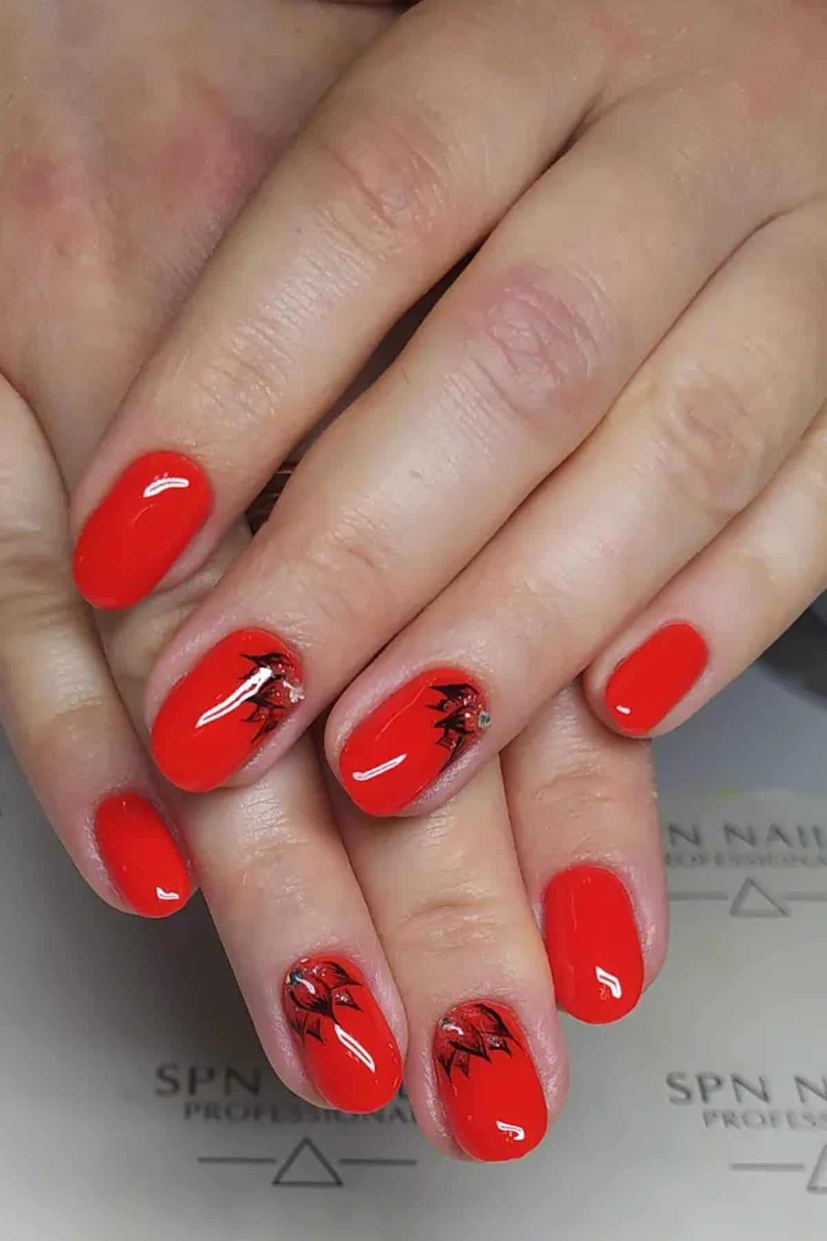 Close-up of hands with short, rounded nails painted bright red; two nails feature black and silver leaf-like nail art designs.