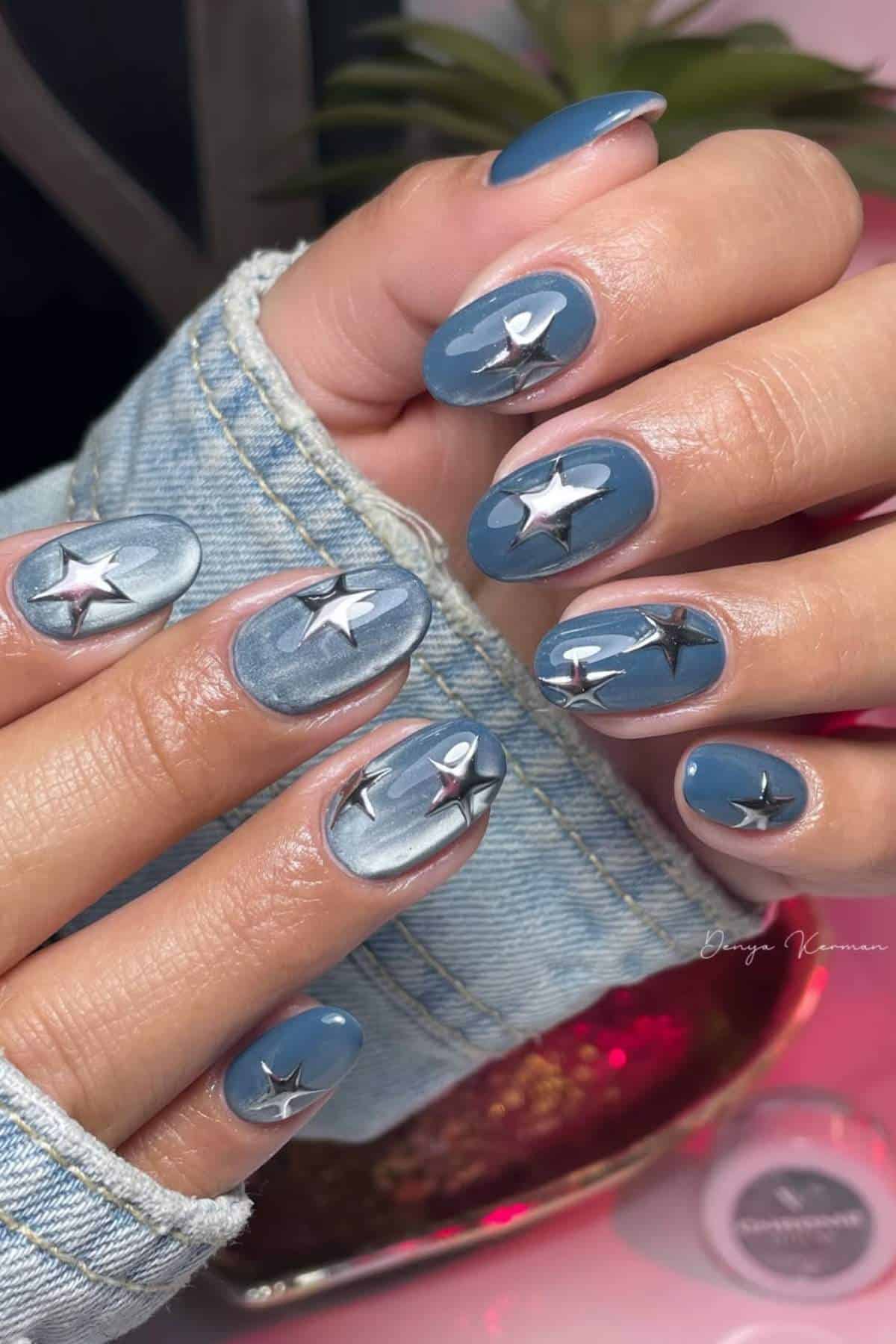 Hands with blue manicured nails featuring silver star and rocket designs, with a denim jacket sleeve visible in the background.