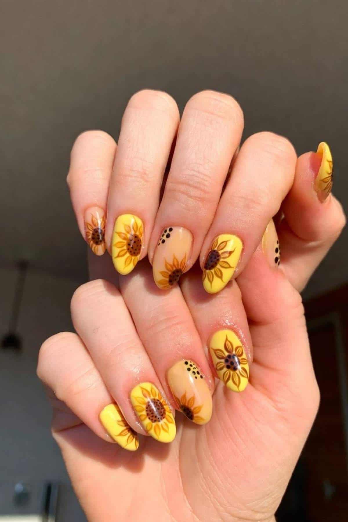 A hand with long, almond-shaped nails painted yellow and decorated with sunflower designs and brown dot accents.