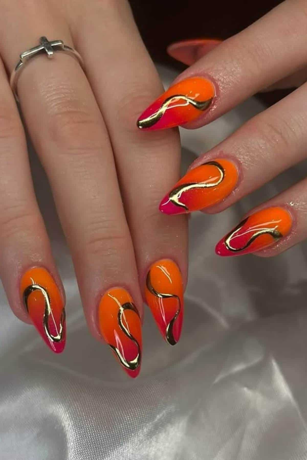 Hand with almond-shaped nails painted in orange and pink with abstract gold and black swirl designs.
