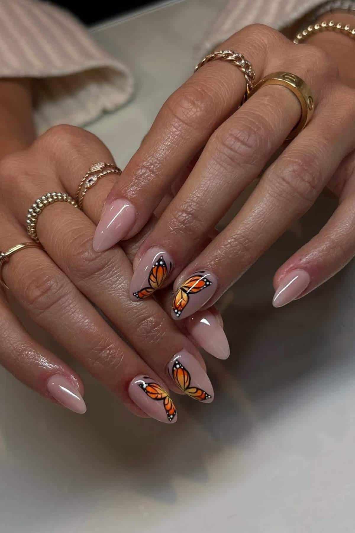 Hands with almond-shaped nails; most are painted nude, while the ring and middle fingernails feature detailed orange and black butterfly wing designs.