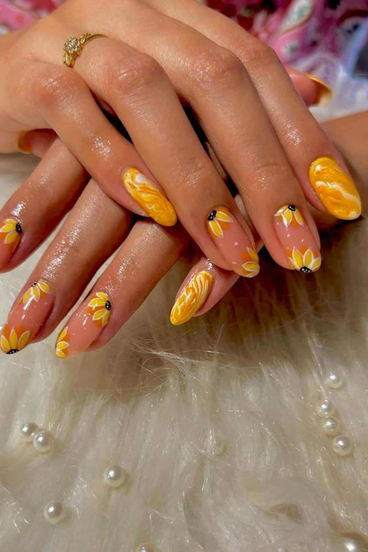 Hands with long almond-shaped nails featuring yellow and white daisy flower designs and marbled yellow patterns, resting on a fuzzy white surface with scattered pearls.