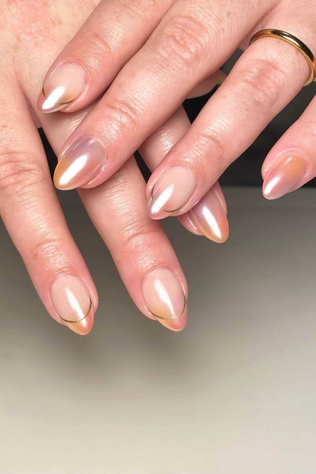 A close-up of hands with almond-shaped nails featuring a glossy nude and gold ombre manicure.