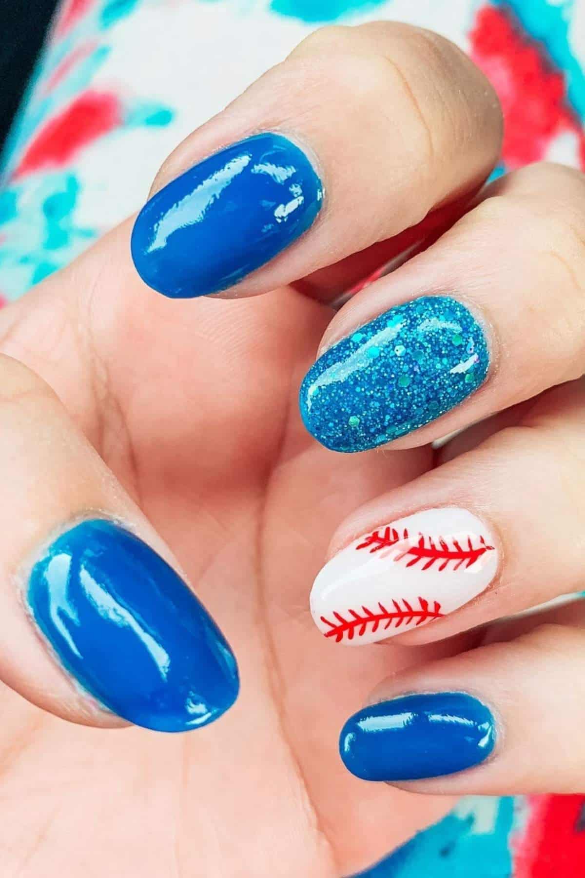 A hand with blue and glittery blue nail polish; the ring fingernail is painted white with red lines resembling a baseball.