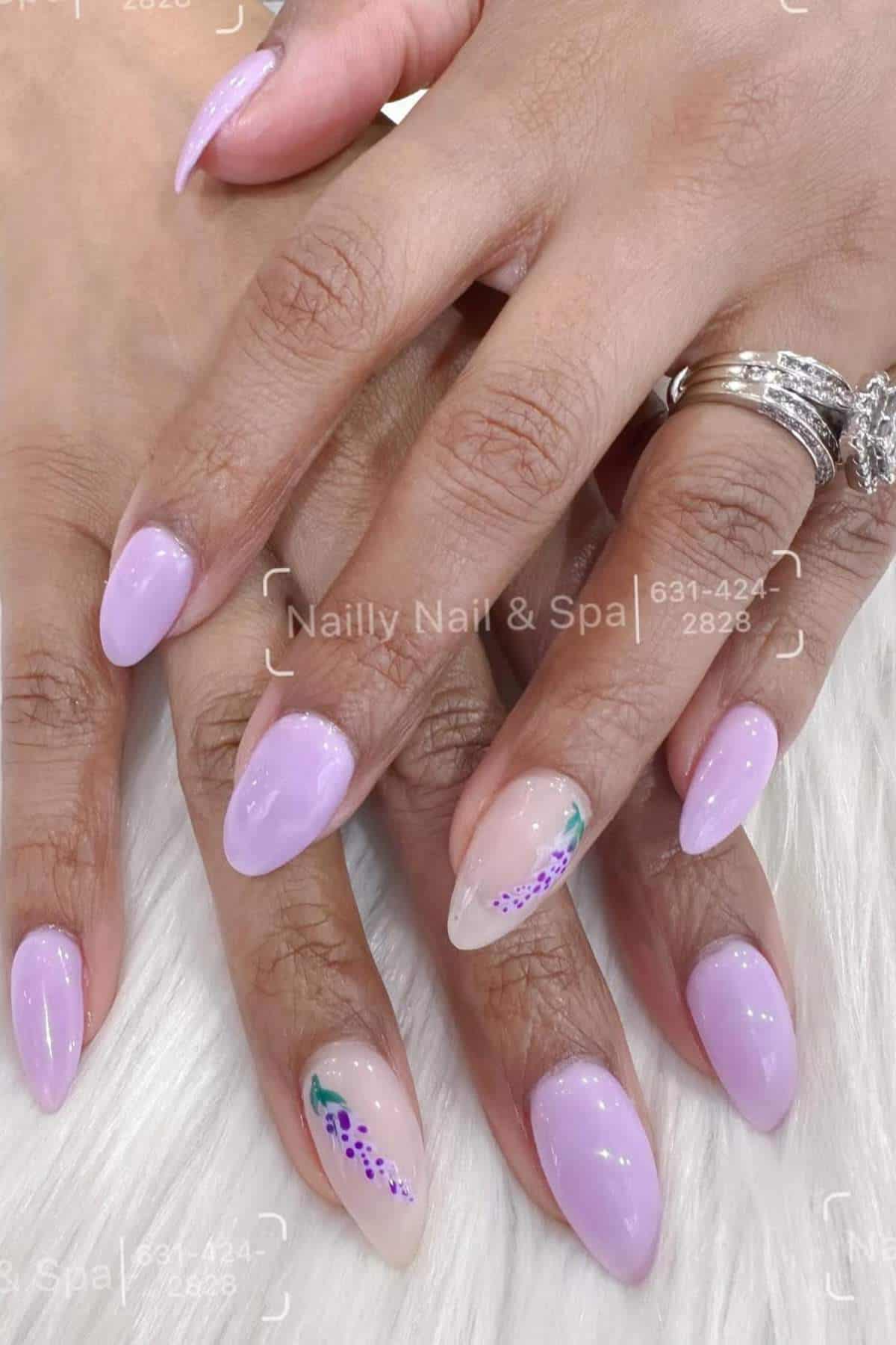 Close-up of hands with oval-shaped, pale lavender nails featuring two accent nails with purple floral designs; person wears rings on two fingers.