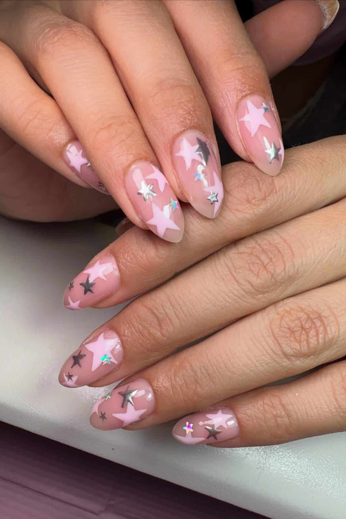 Hands with almond-shaped nails featuring a light pink base and decorated with silver and pink star-shaped decals.