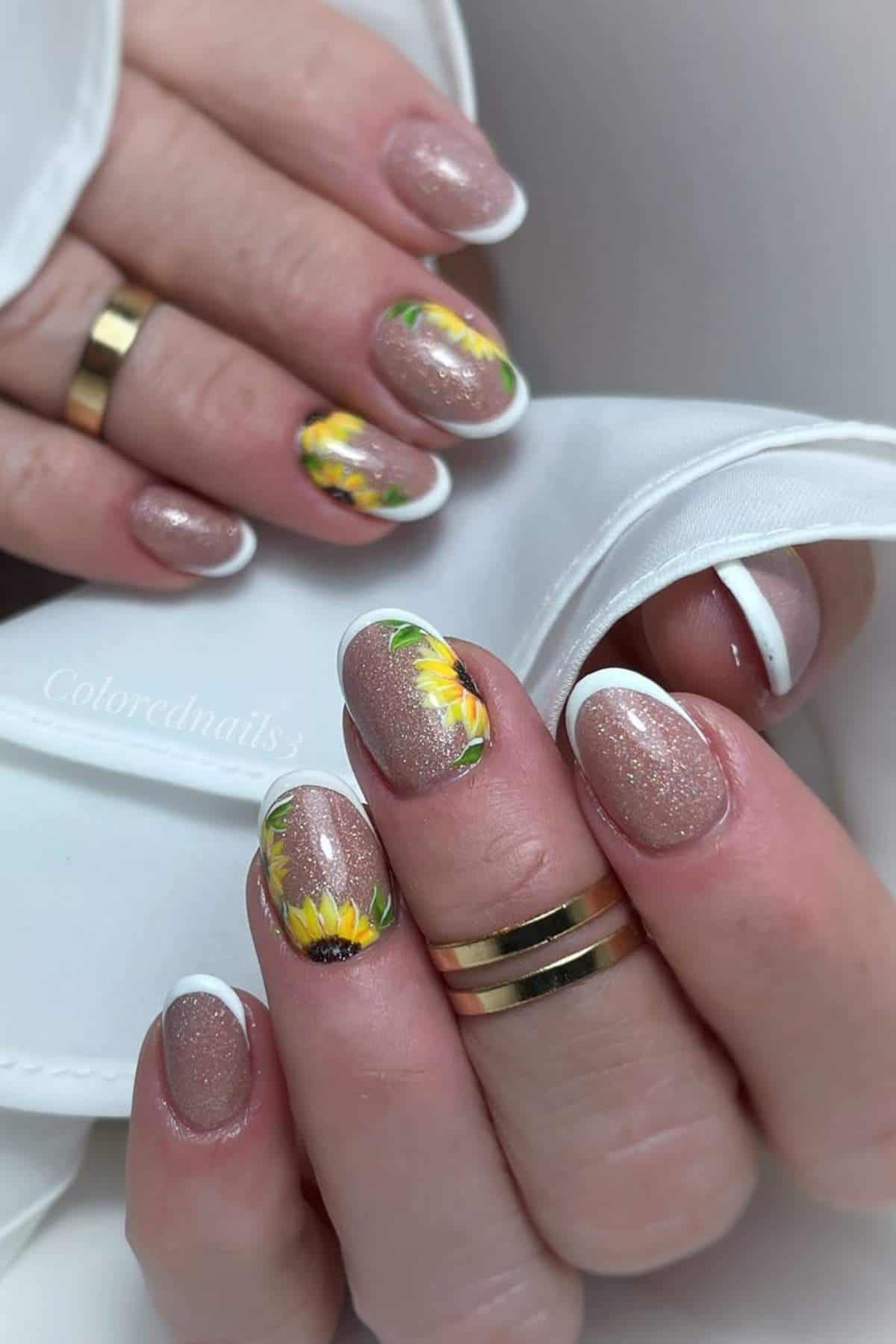 A hand with nude glittery nails, white French tips, and sunflower designs on two accent nails, holding a white fabric. Gold rings are worn on two fingers.
