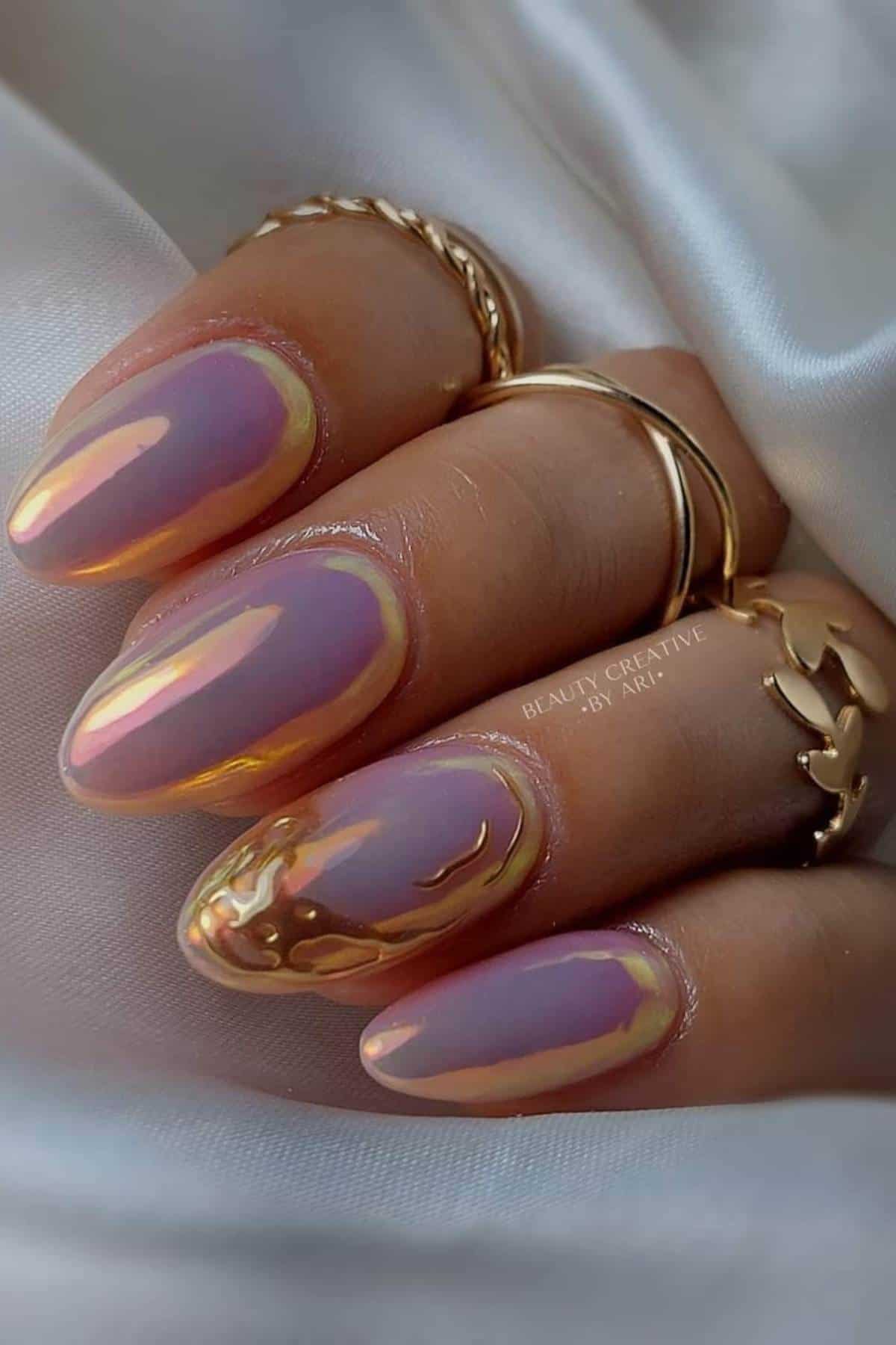 A hand with long, almond-shaped nails painted in an iridescent lavender polish with gold chrome accents, wearing three gold rings, against a satin fabric background.