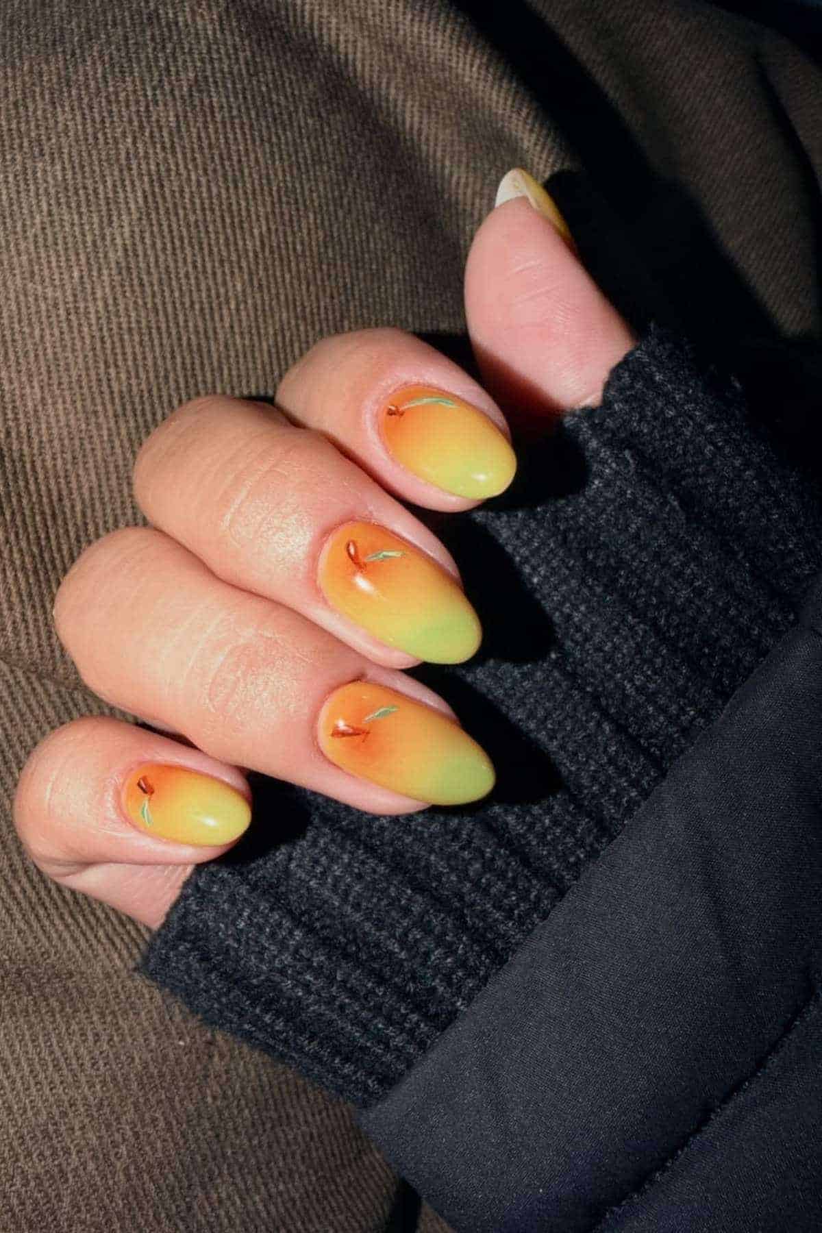 A hand with almond-shaped nails painted in a yellow-to-green ombre finish with small orange flame designs, shown against a dark textured fabric background.