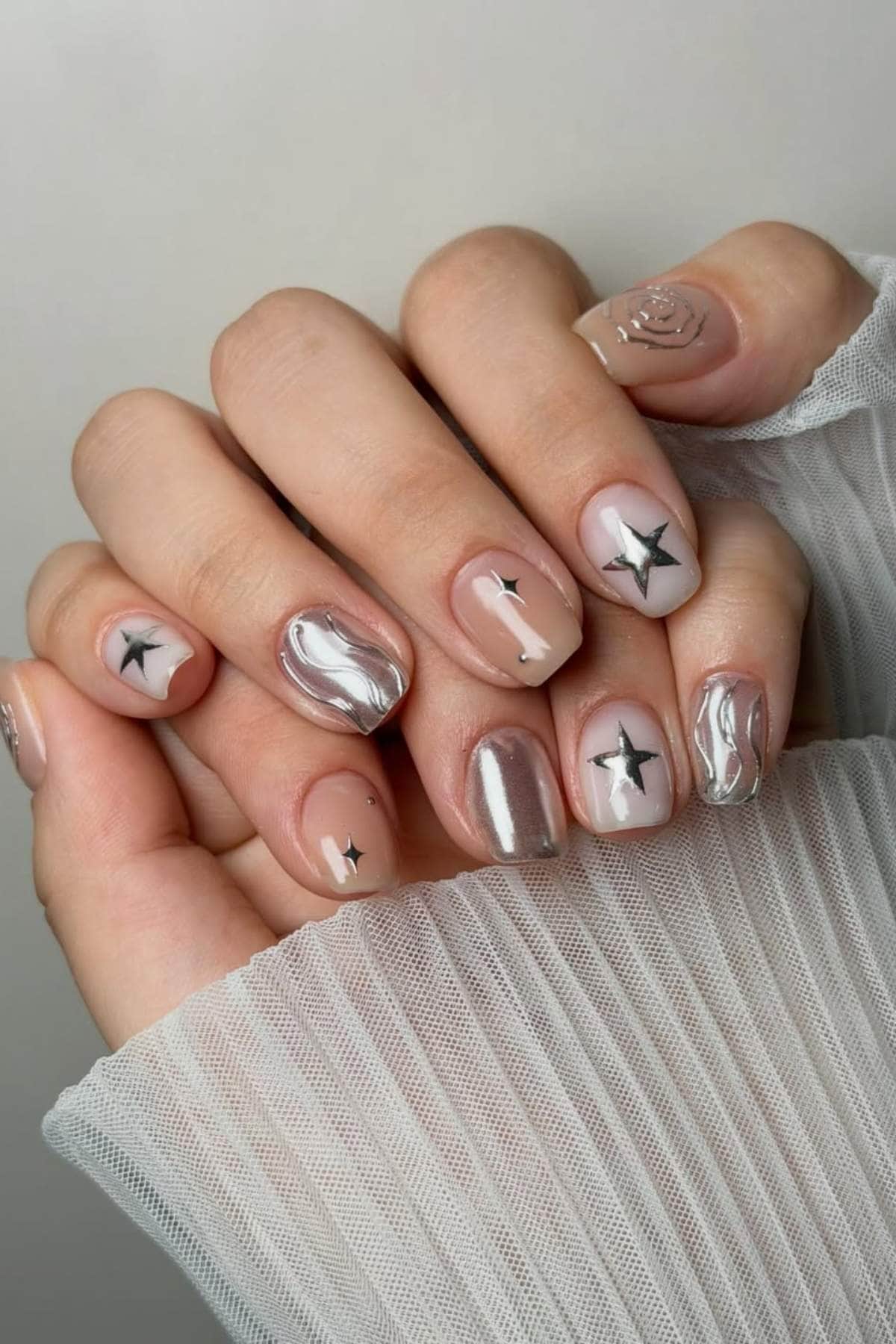 A hand with short, squared nails painted in a mix of pearly silver, nude, and translucent shades, featuring black star designs and metallic swirls.