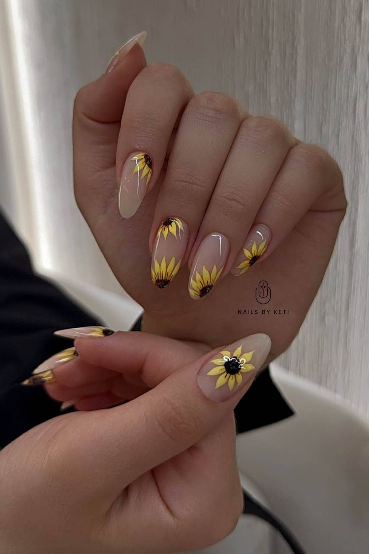 A hand with long, almond-shaped nails featuring sunflower nail art on a nude base, displayed against a neutral background.