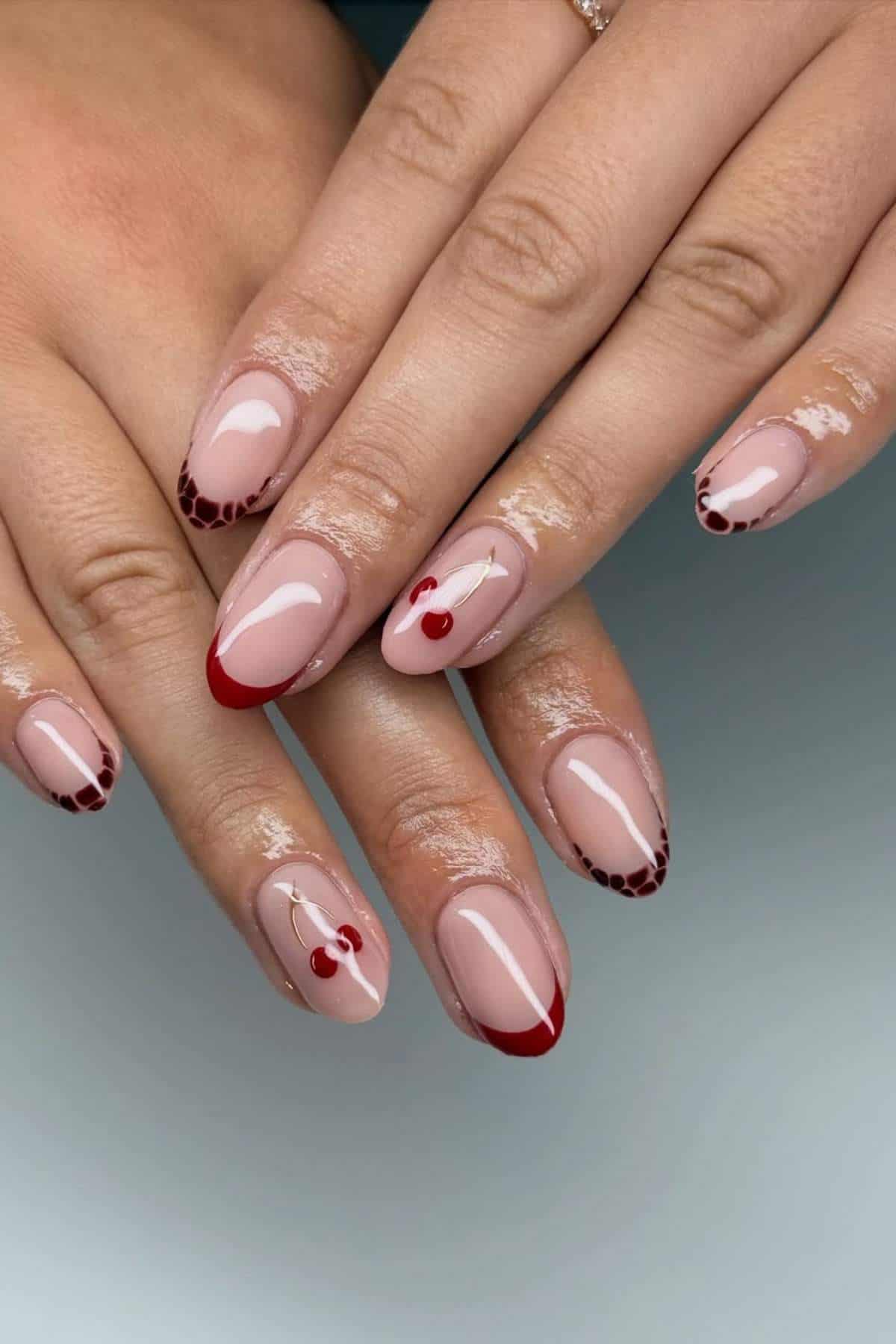A hand with almond-shaped nails featuring nude polish, red French tips, cherry designs, and brown patterned accents near the tips.