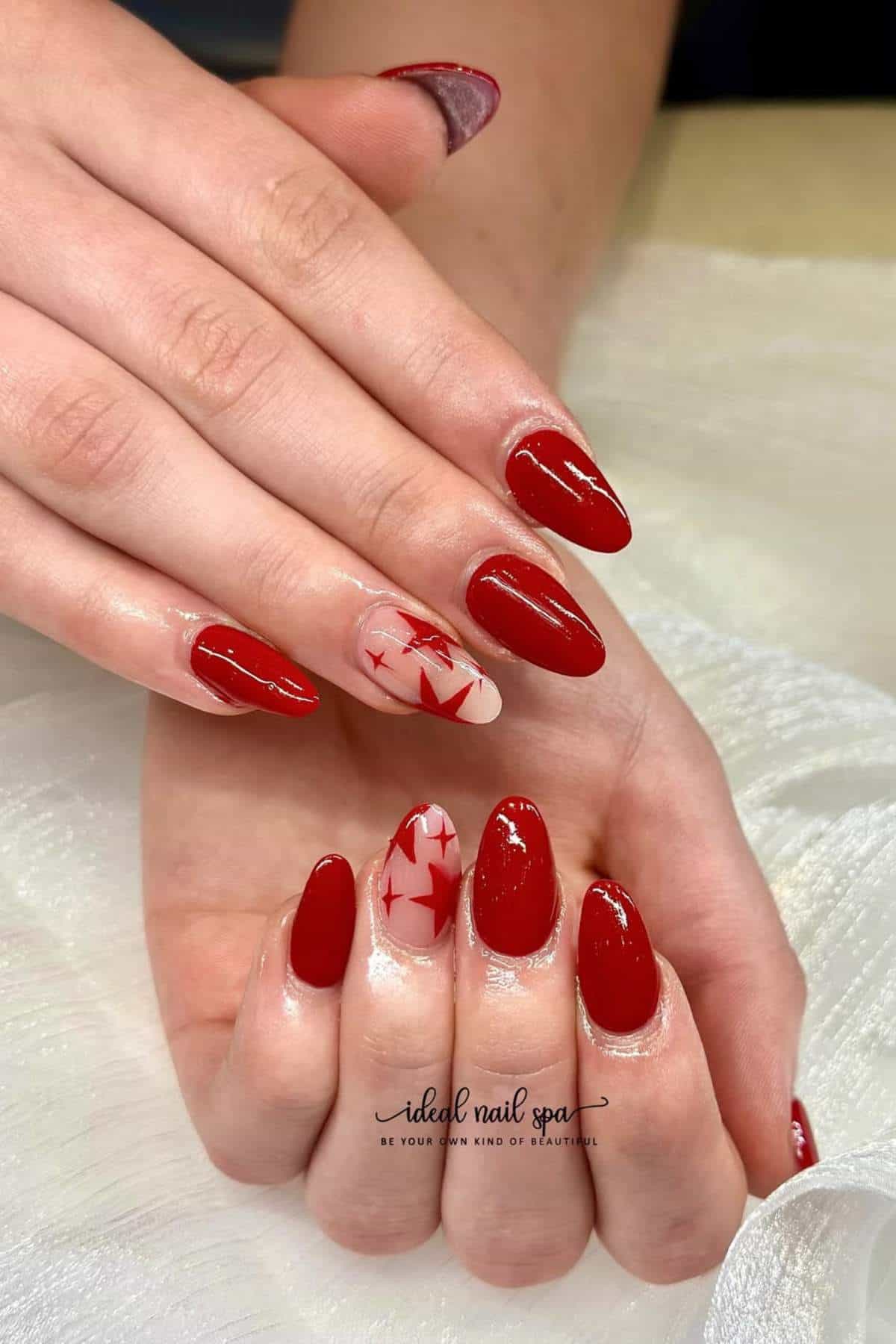Hands with almond-shaped nails, most painted glossy red; one accent nail on each hand is nude with red geometric star designs.