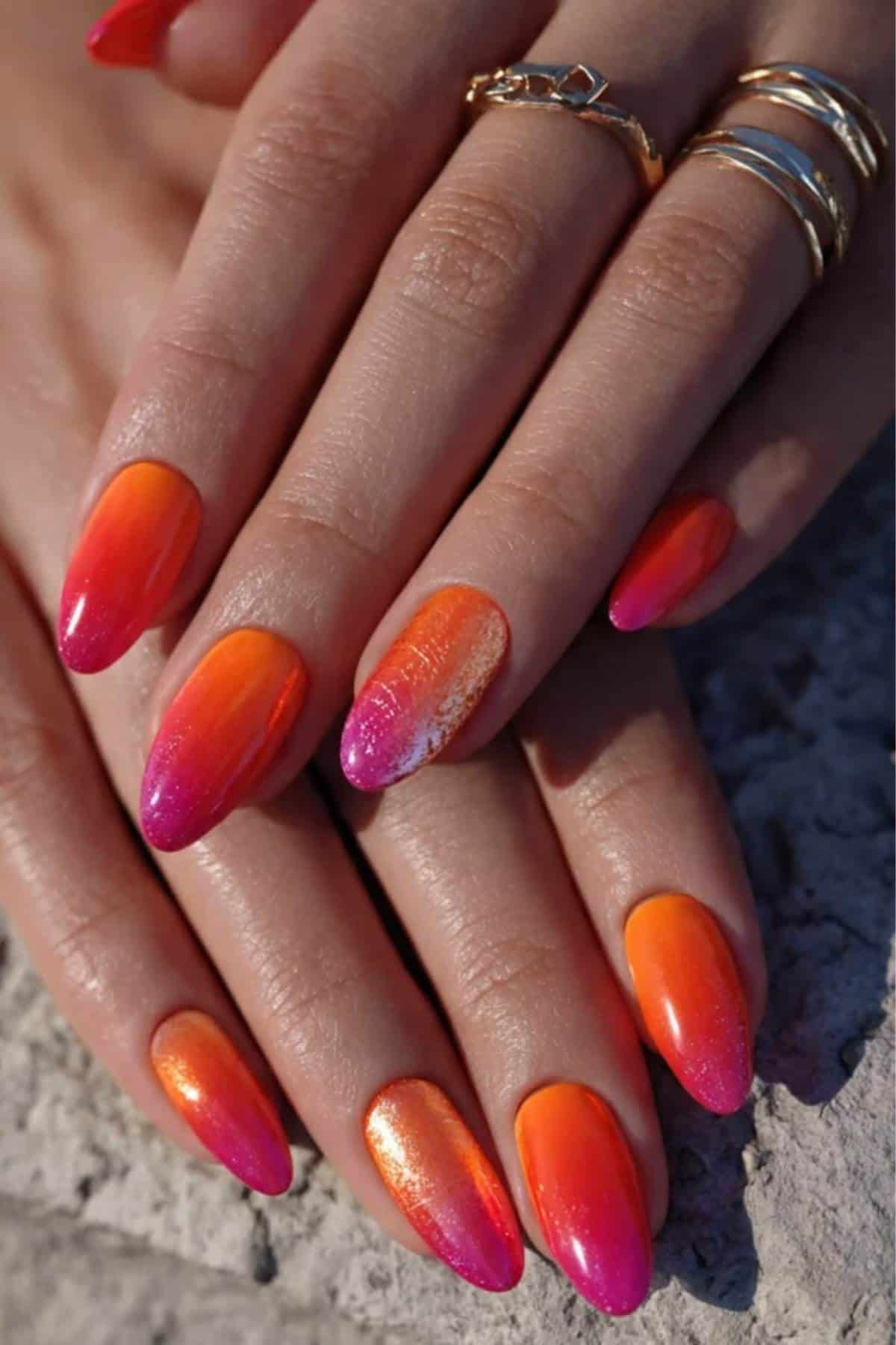 A close-up of hands with long, almond-shaped nails featuring an orange to pink ombre manicure and gold rings on the fingers.