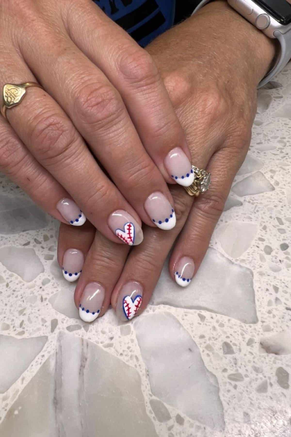 Hands with manicured nails; most nails have white French tips with blue dots, while two feature red baseball stitching designs.