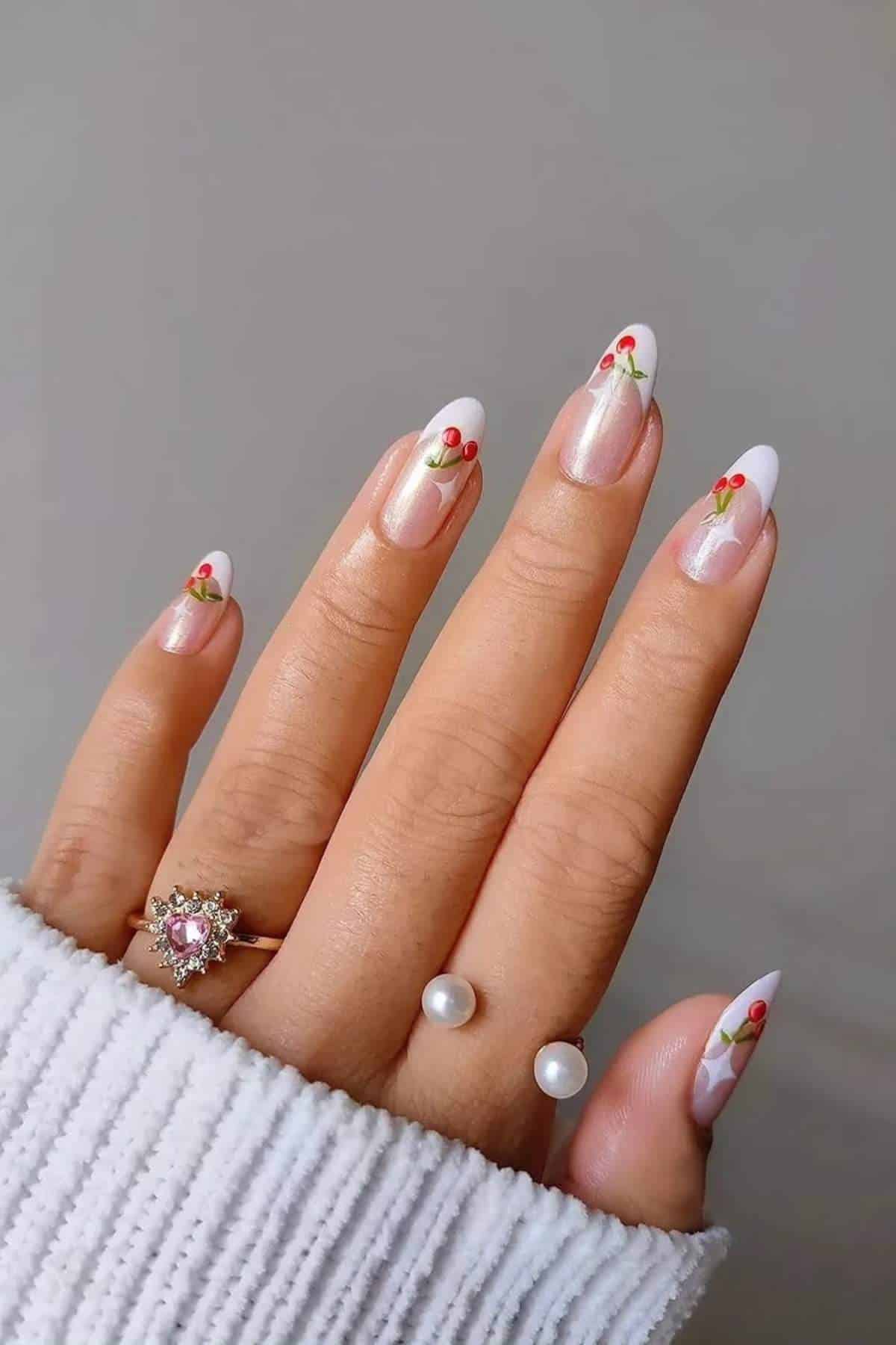 A hand with almond-shaped nails featuring white tips and small red cherry designs, wearing a pearl ring and a gold ring with a pink heart-shaped stone.