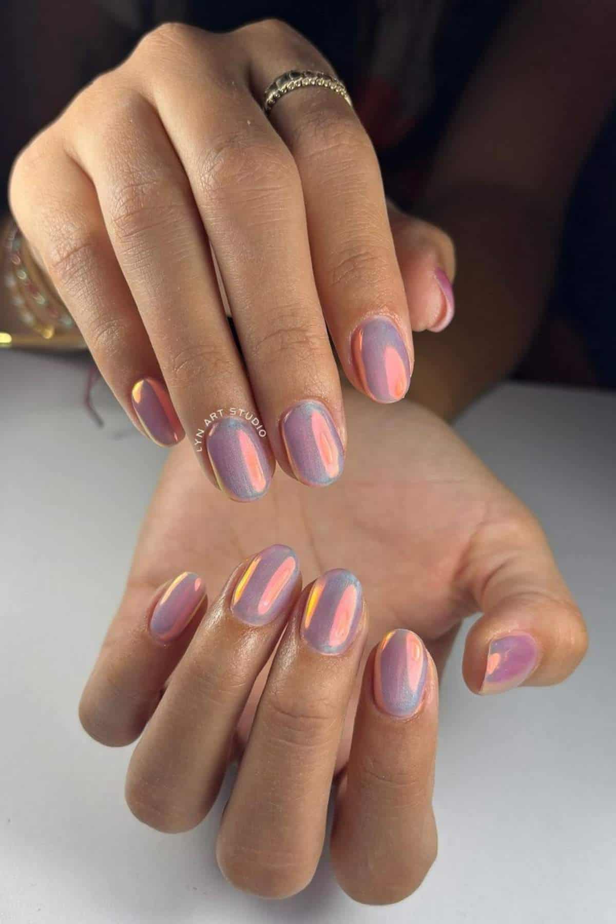 Close-up of hands with short, oval-shaped nails painted in a purple iridescent polish with pink and yellow reflections.