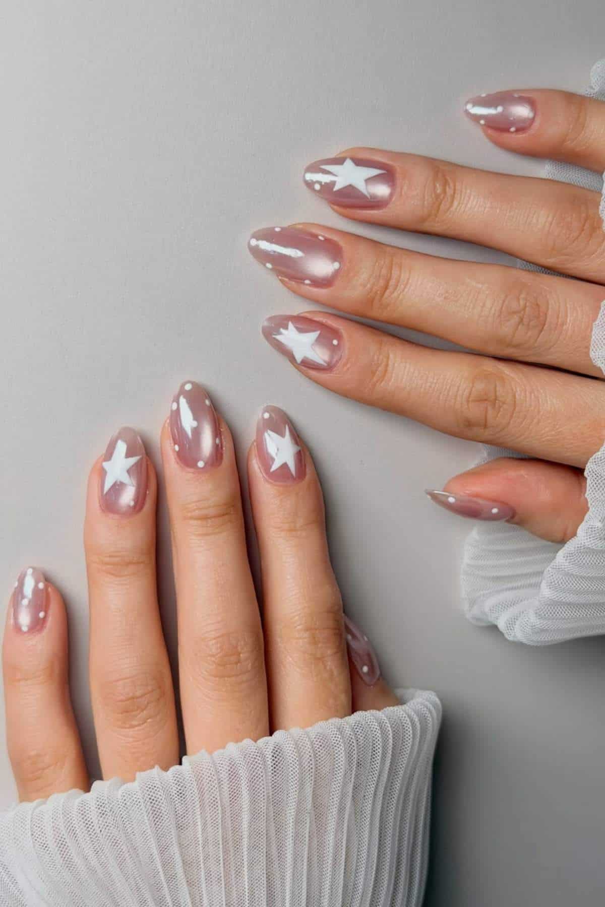 Hands with long, almond-shaped nails painted in a sheer pink polish with white star and dot designs. The person is wearing a white, pleated, sheer fabric top.