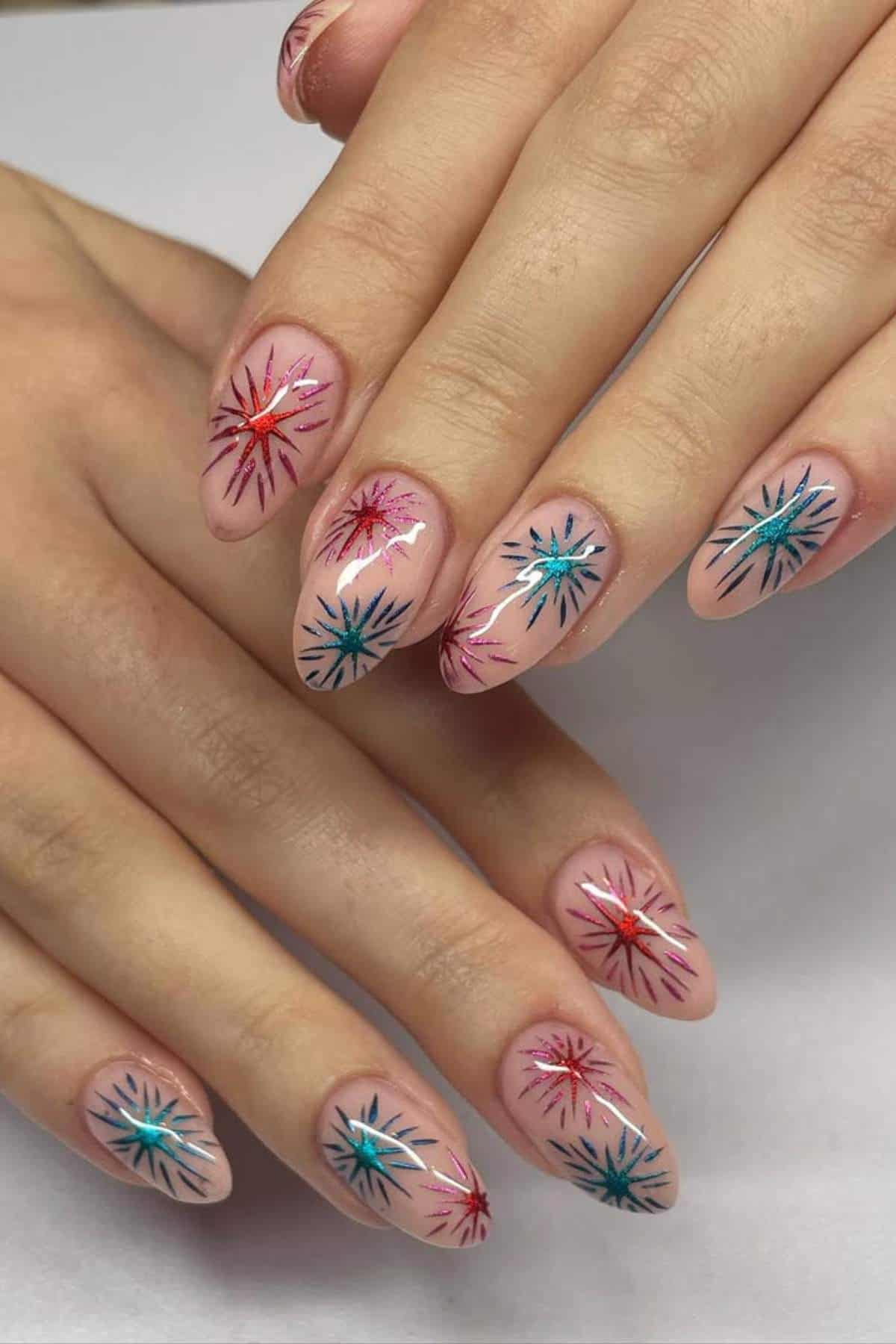 Hands with almond-shaped nails featuring a nude base and colorful starburst designs in red, blue, and teal on each nail.