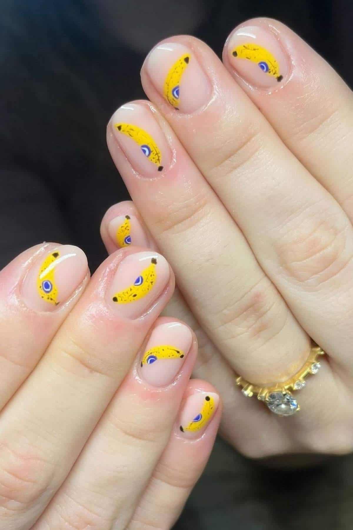 Close-up of hands with short, natural nails featuring yellow banana nail art with blue accents on a clear base.