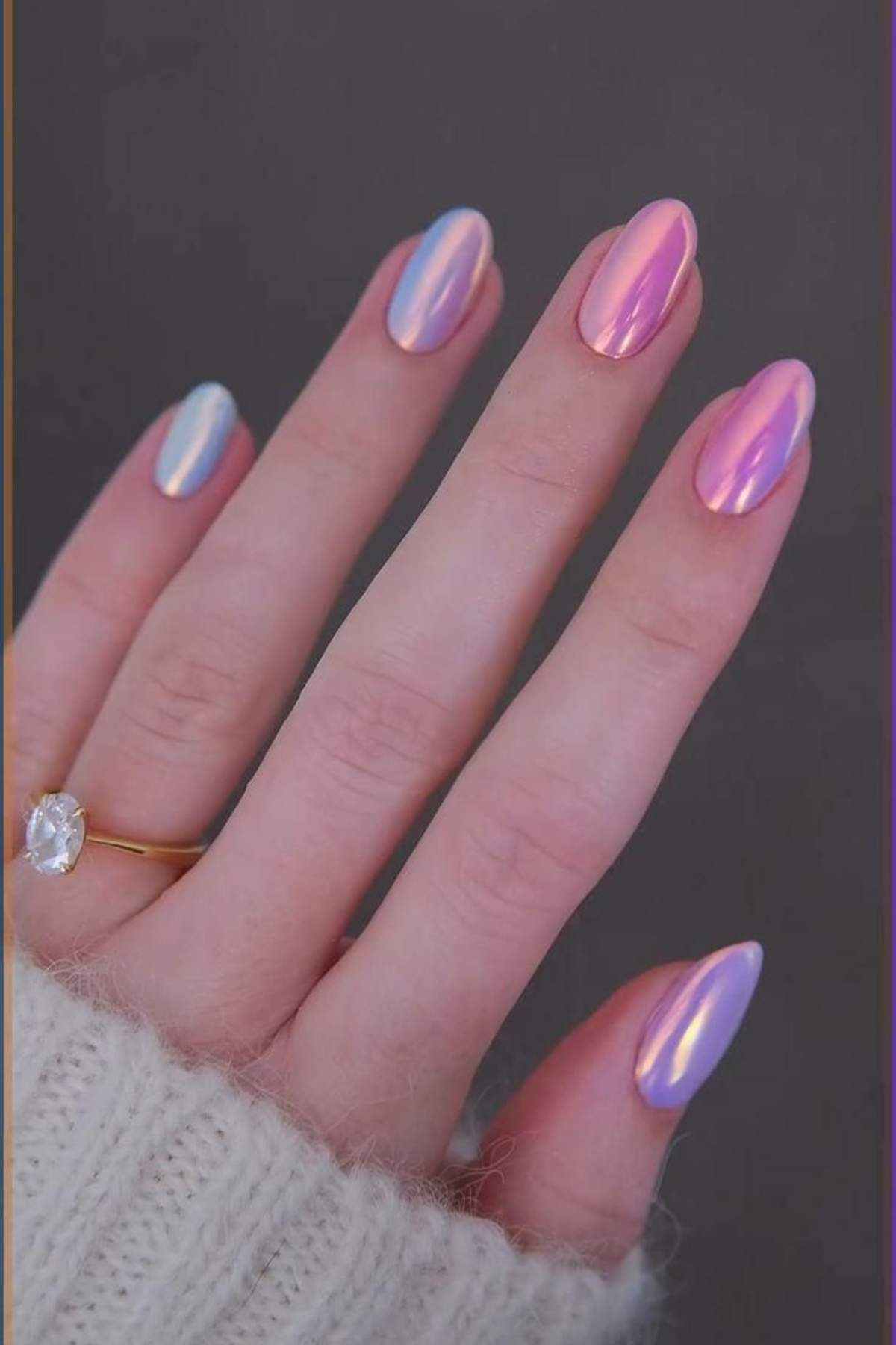 A hand with almond-shaped, iridescent pink and blue chrome nails, wearing a gold ring with a large round stone, and a cream knit sleeve visible.