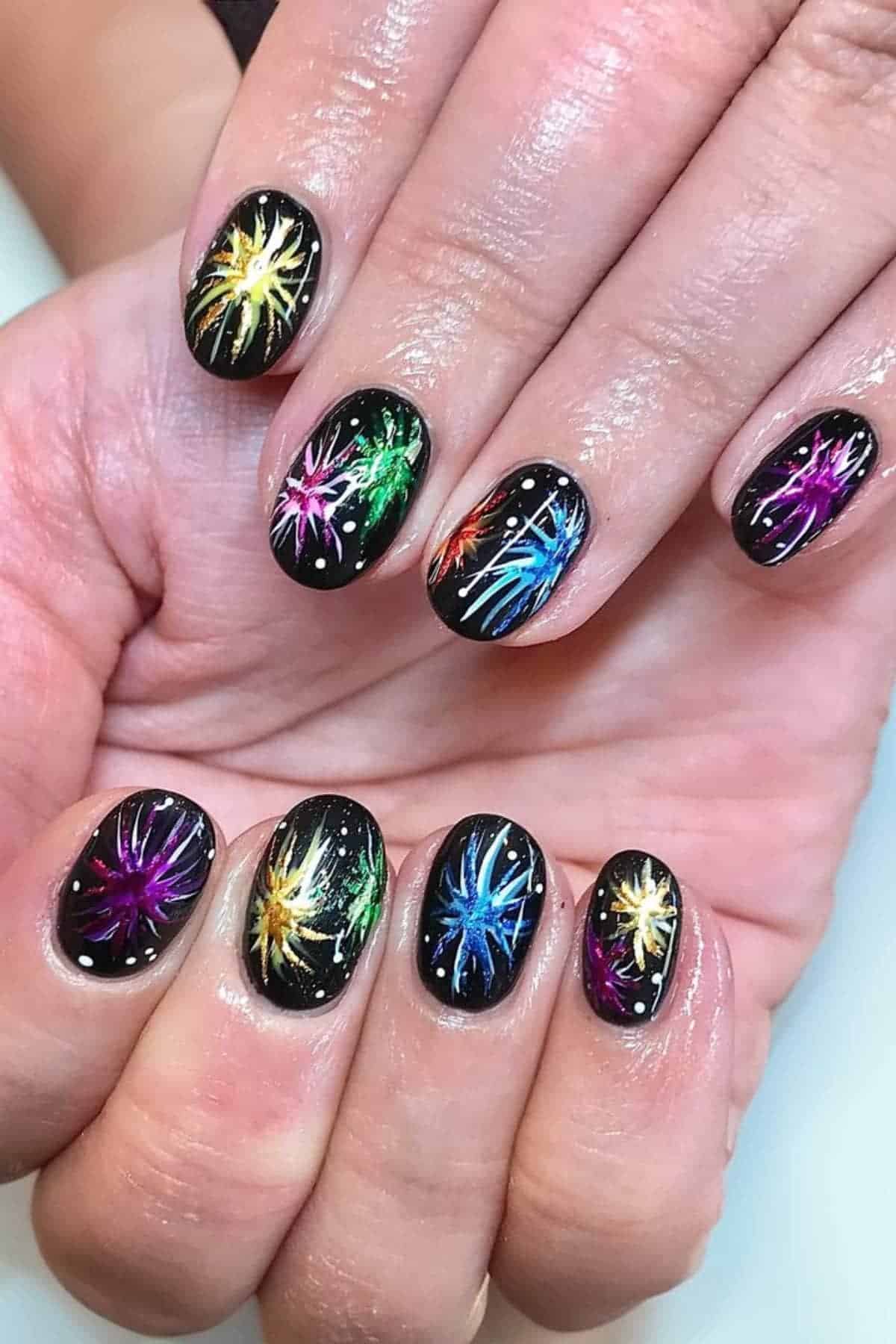 Hands with short, oval nails painted black and decorated with colorful fireworks designs and small white dots, resembling stars, on each nail.