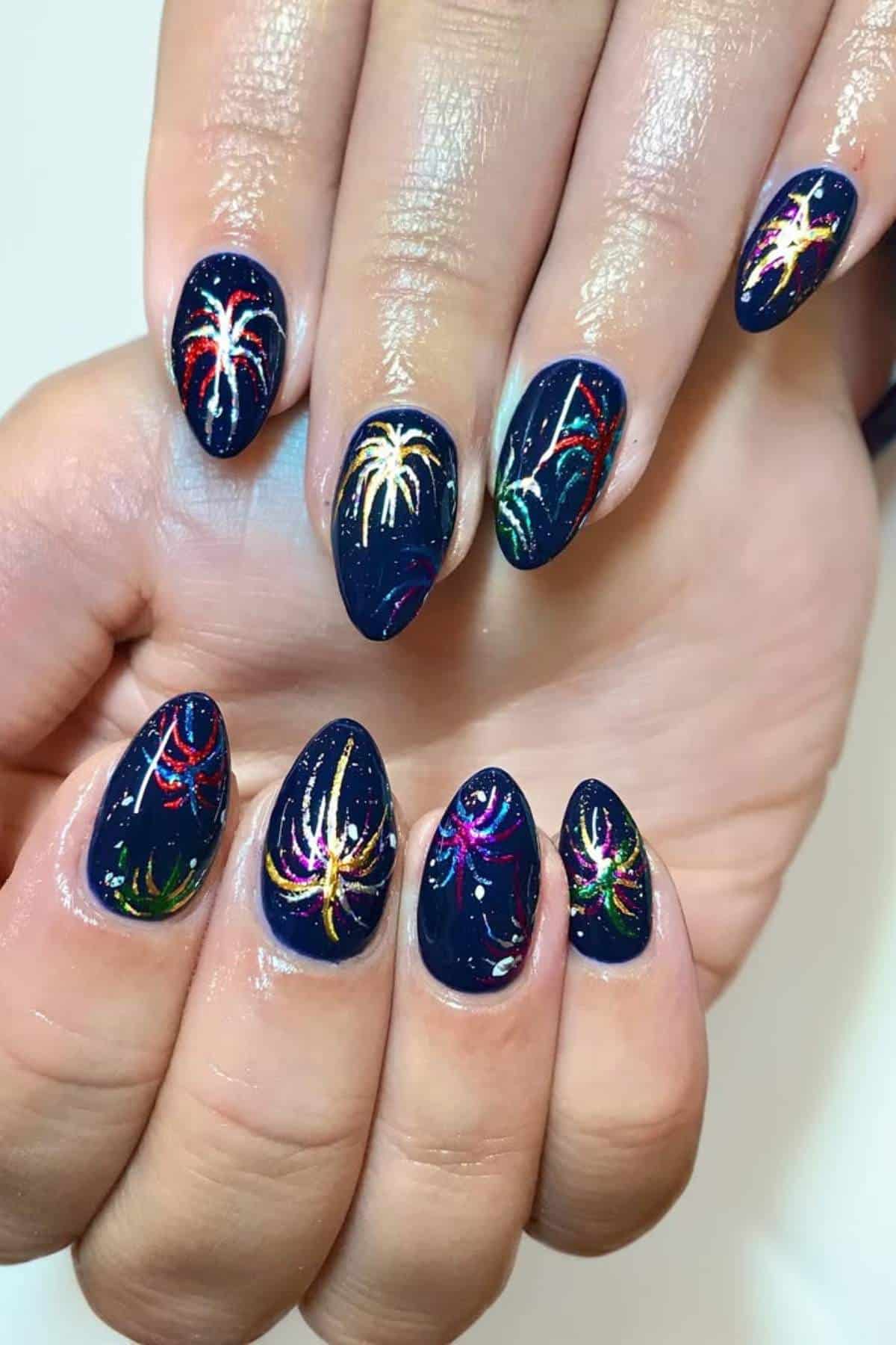 Hands with almond-shaped nails painted dark blue, featuring colorful firework-like designs in red, yellow, green, purple, and white on each nail.