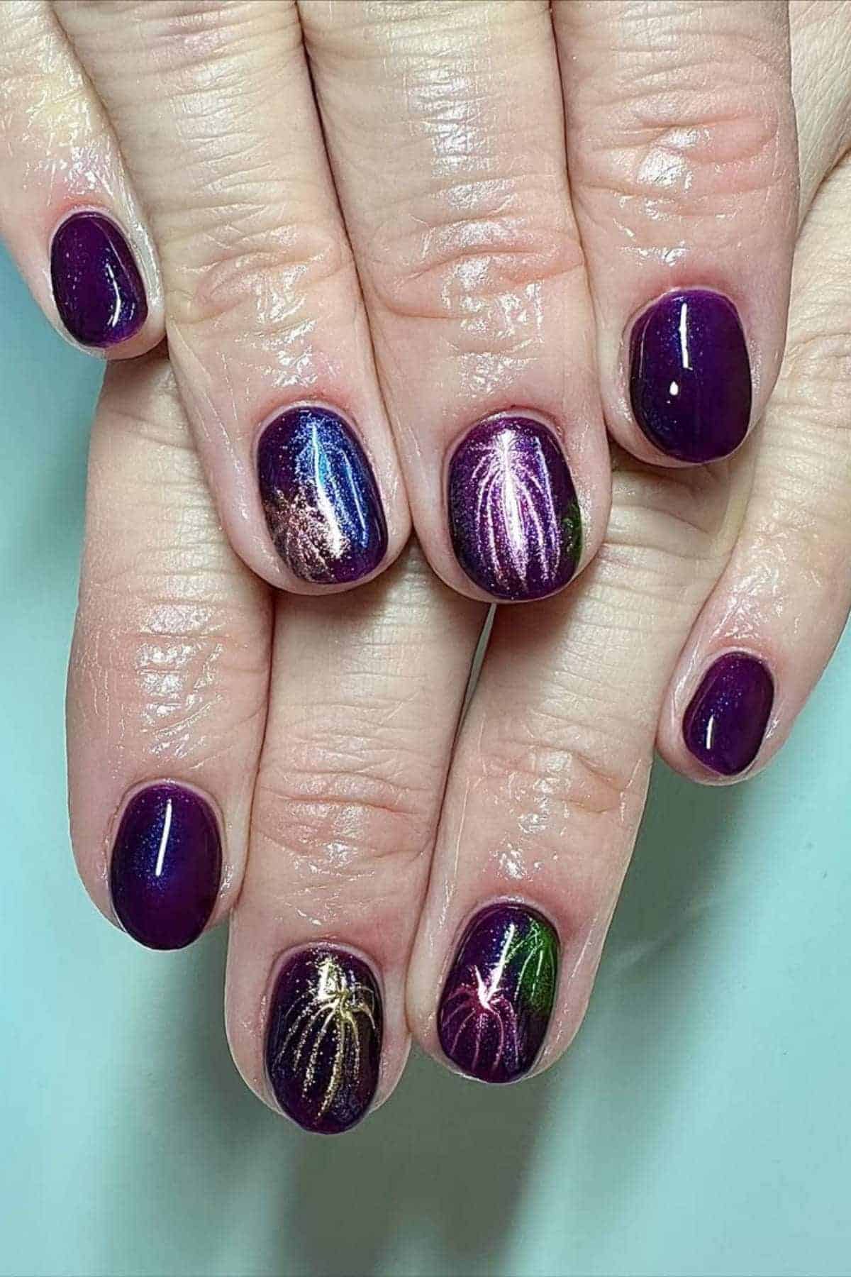 Hands with short, rounded nails painted dark purple with a glossy finish; some nails feature multicolored firework-style designs.