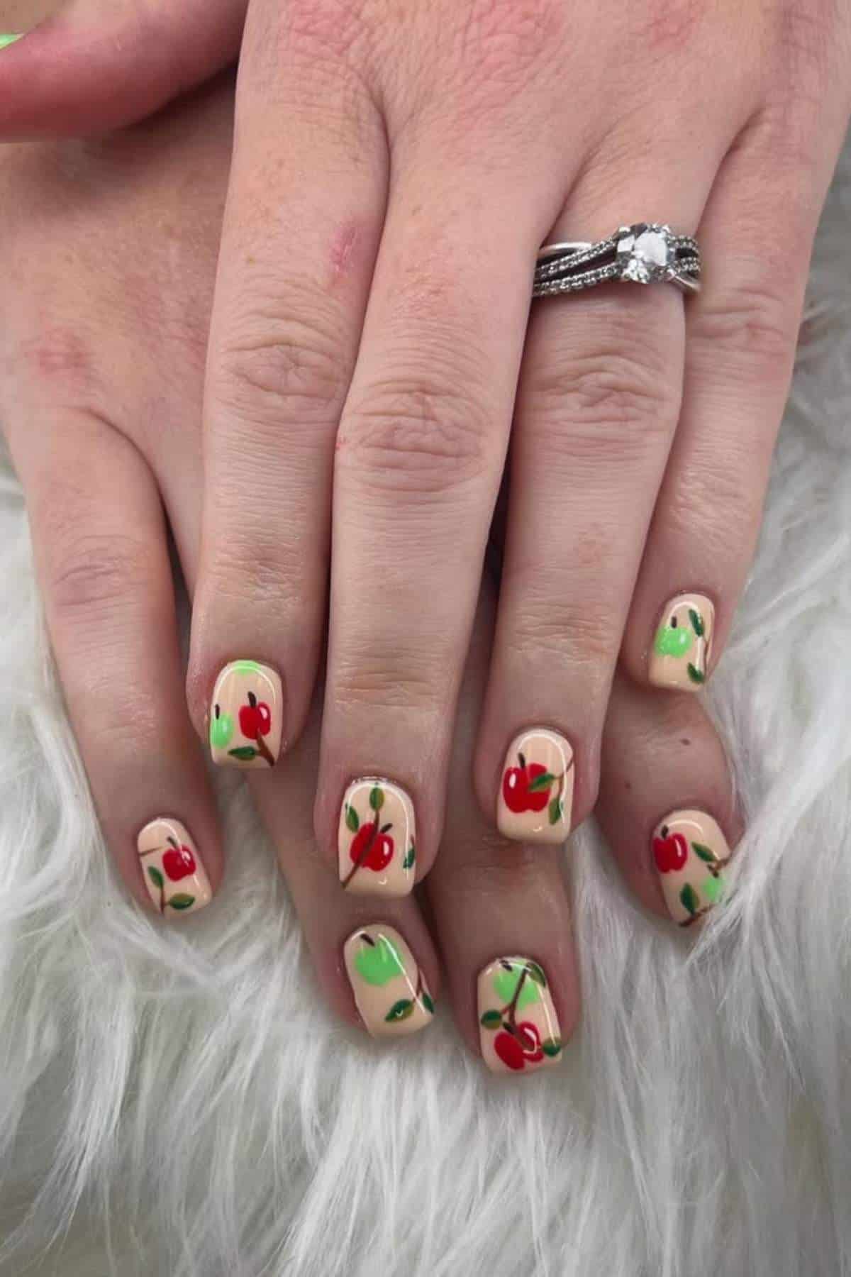 Hands with short nails painted with a nude base and decorated with red cherry nail art; one hand has a diamond ring on the ring finger.