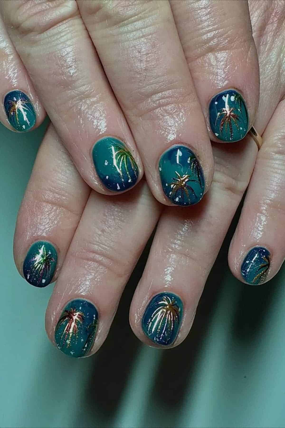 Close-up of hands with teal nail polish featuring detailed hand-painted firework designs on each fingernail.