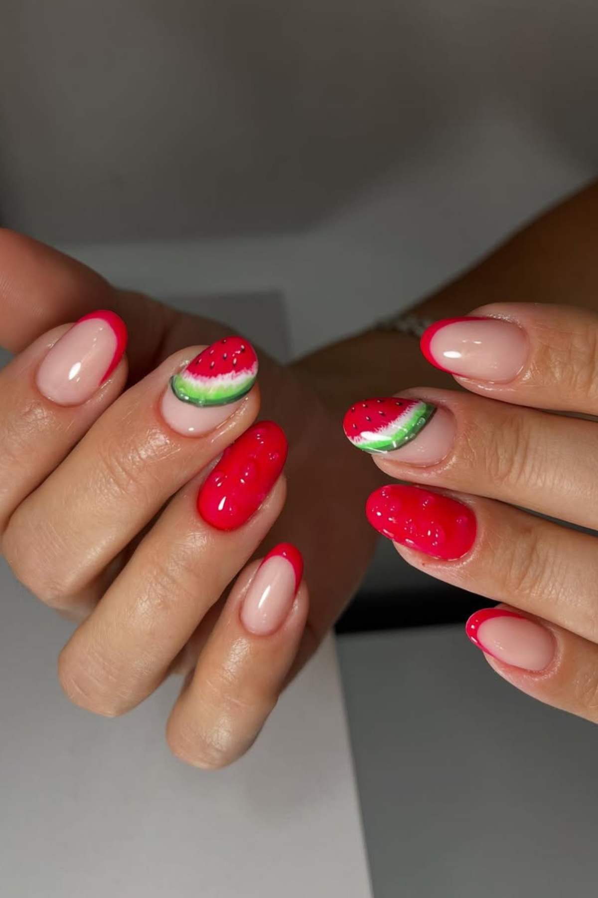 Hands with manicured nails featuring a watermelon design on two nails, bright red polish on others, and a nude base with red tips on some.