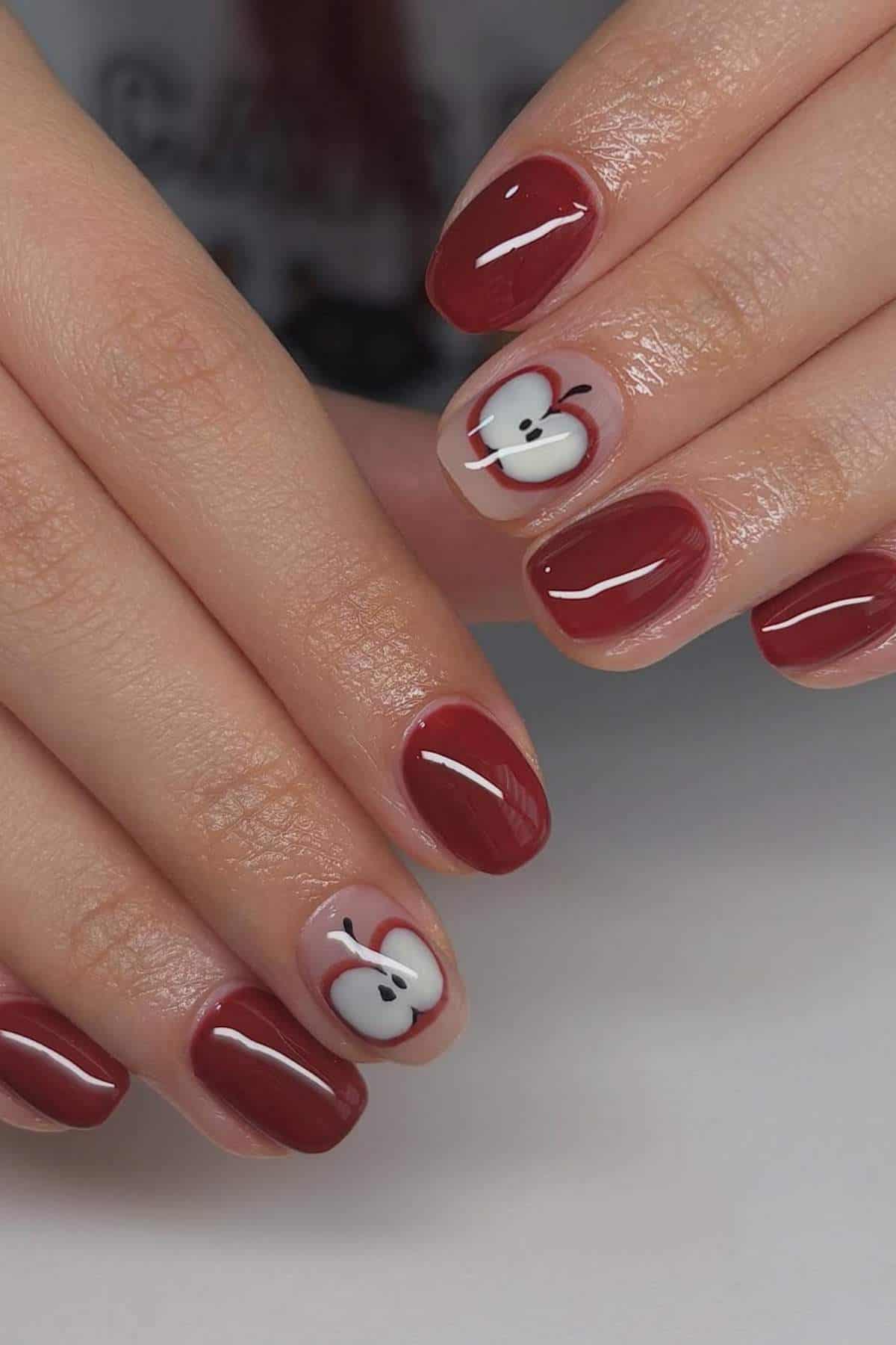 A close-up of hands with glossy dark red nail polish; two accent nails feature a design of a sliced apple with seeds and a brown outline.