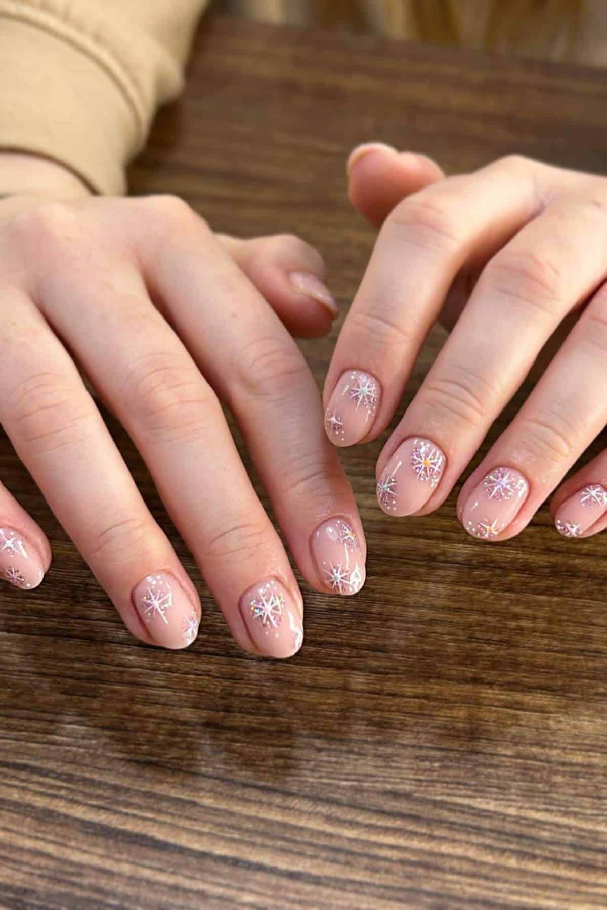 Two hands with short, oval nails painted nude and decorated with white snowflake designs rest on a wooden surface.