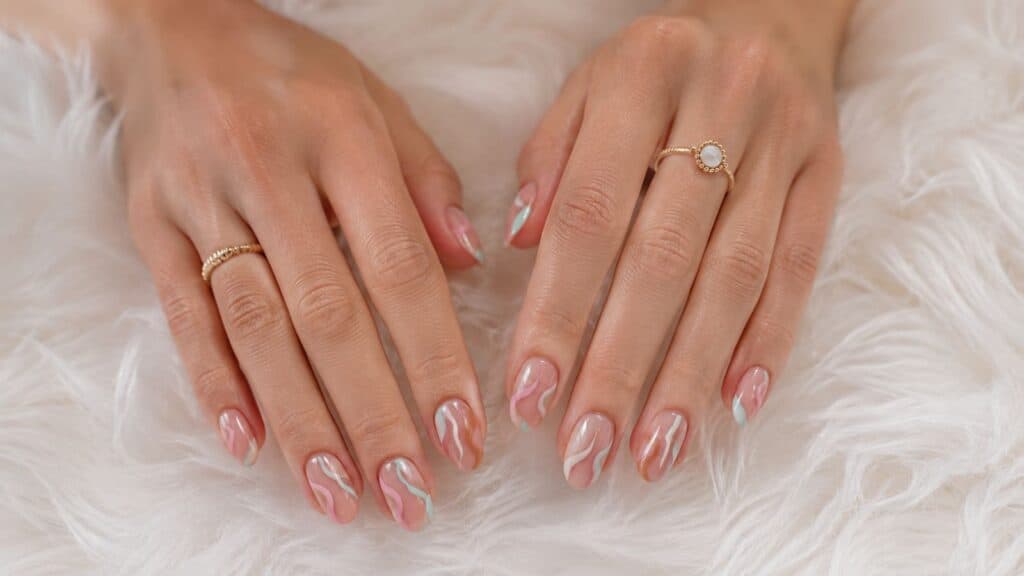Two hands with manicured nails featuring an abstract pink and white swirl design, resting on a white fluffy surface. The person wears gold rings on both hands.