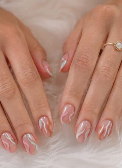 Two hands with manicured nails featuring an abstract pink and white swirl design, resting on a white fluffy surface. The person wears gold rings on both hands.