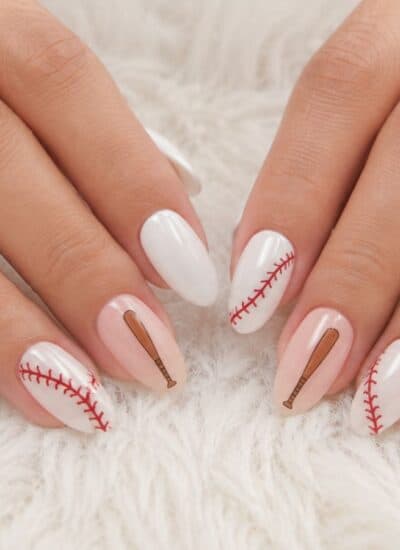 Hands with white manicured nails featuring baseball seam and bat designs, resting on a soft white textured surface.